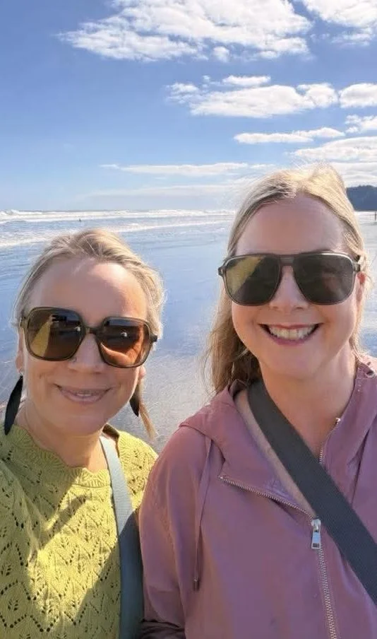 Two women smiling at the beach, both wearing sunglasses, with waves and a blue sky with clouds in the background.
