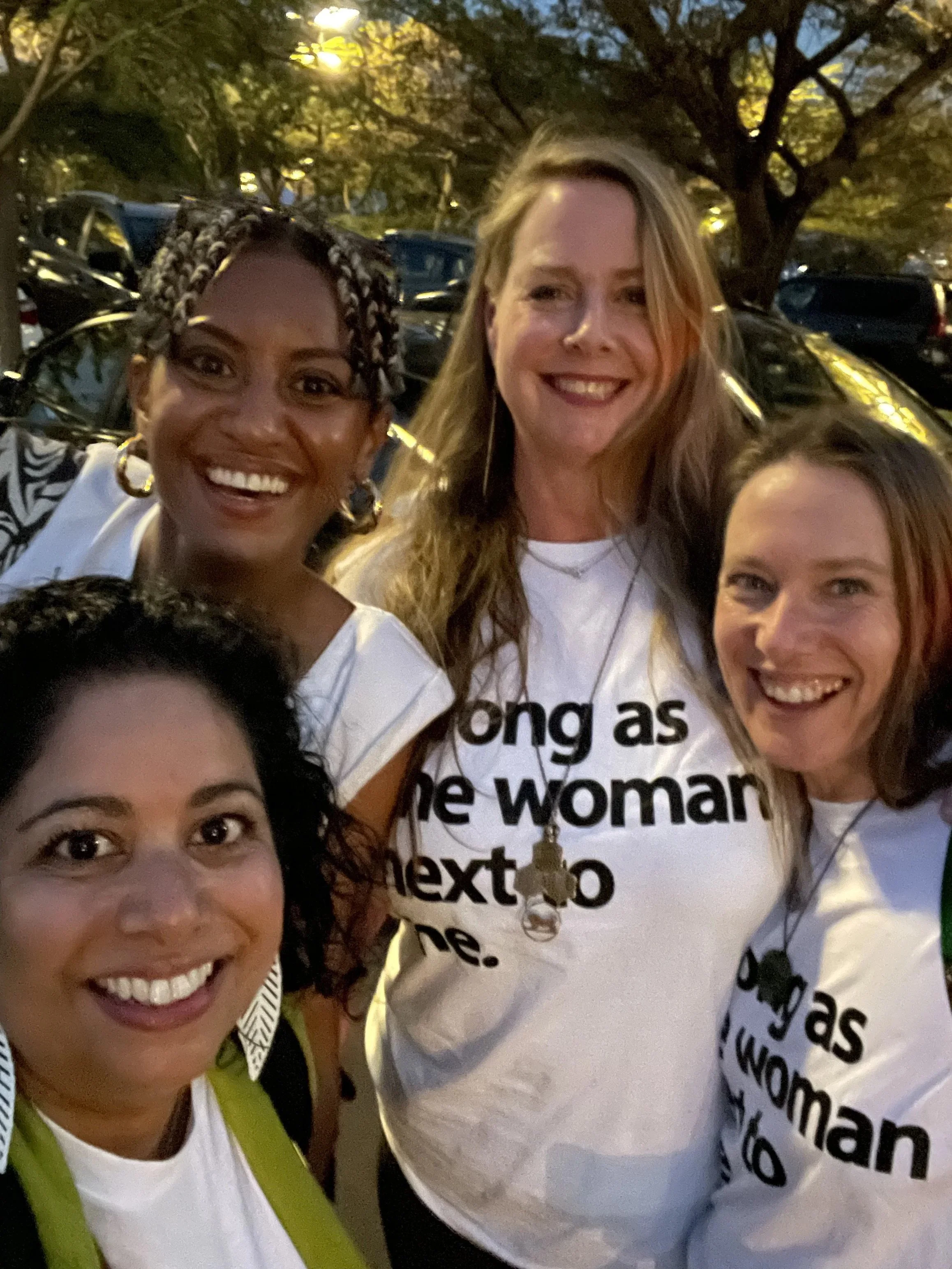 A group of women smiling and taking a selfie outdoors during the evening with trees and parked cars in the background. One woman is wearing a white shirt with black text and a necklace with keys. Another woman has braids and large hoop earrings.