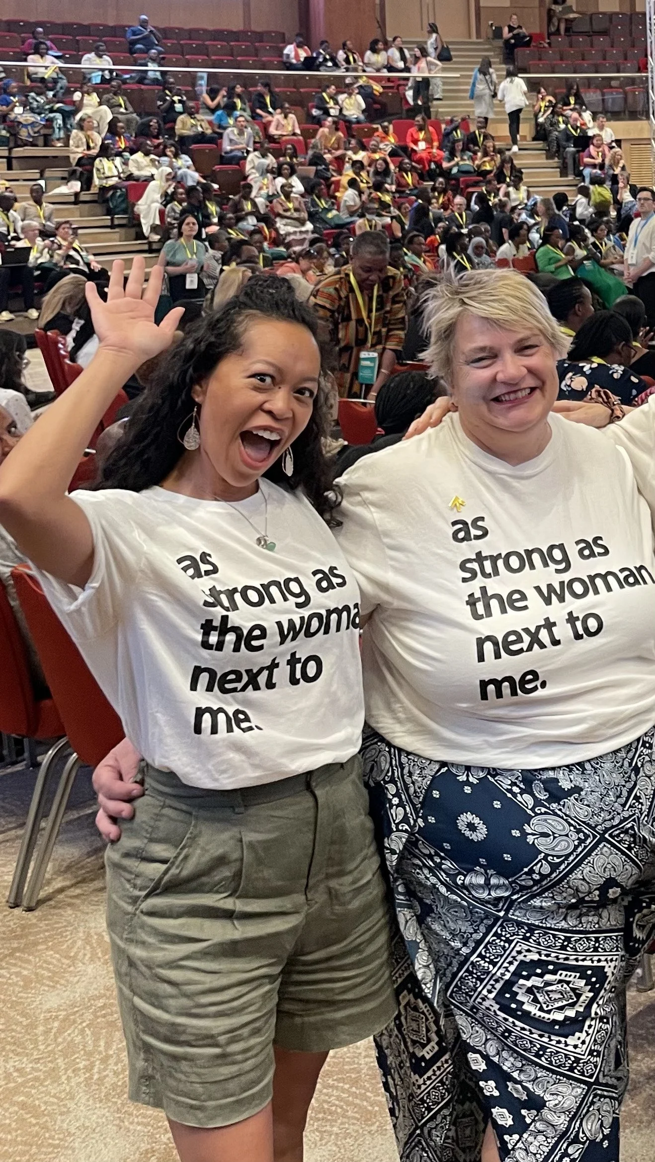 Two women smiling and posing at a large indoor event. Both wearing matching white t-shirts that say 'as strong as the woman next to me.' The woman on the left has long curly hair, earrings, and khaki shorts. The woman on the right has short blonde hair and patterned skirt. The background shows many people seated in stadium-style seating, some wearing same-style lanyards.