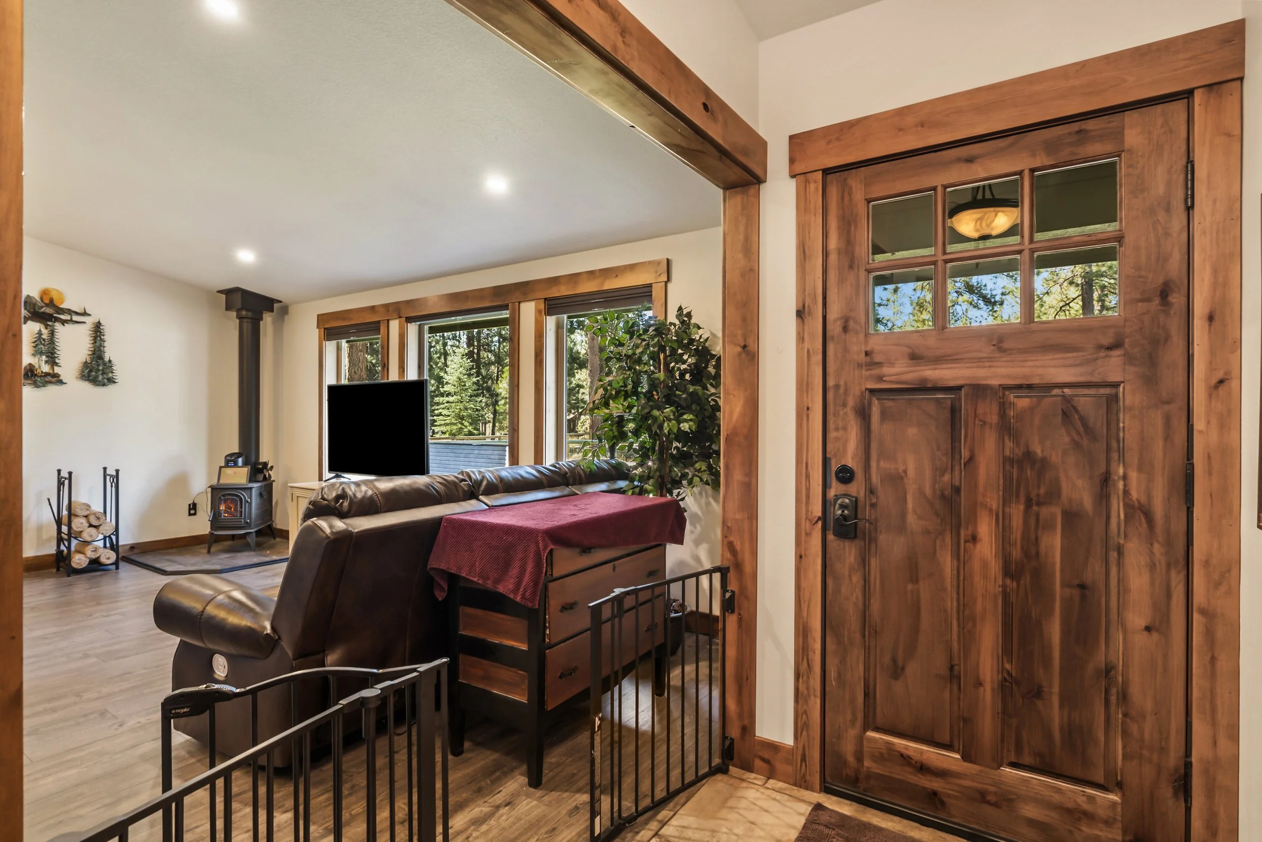 Interior view of a living room with wooden trim and large windows showing trees outside. There is a brown leather sofa, a small black fireplace, and stacks of firewood. A wood door with glass panes is in the foreground.