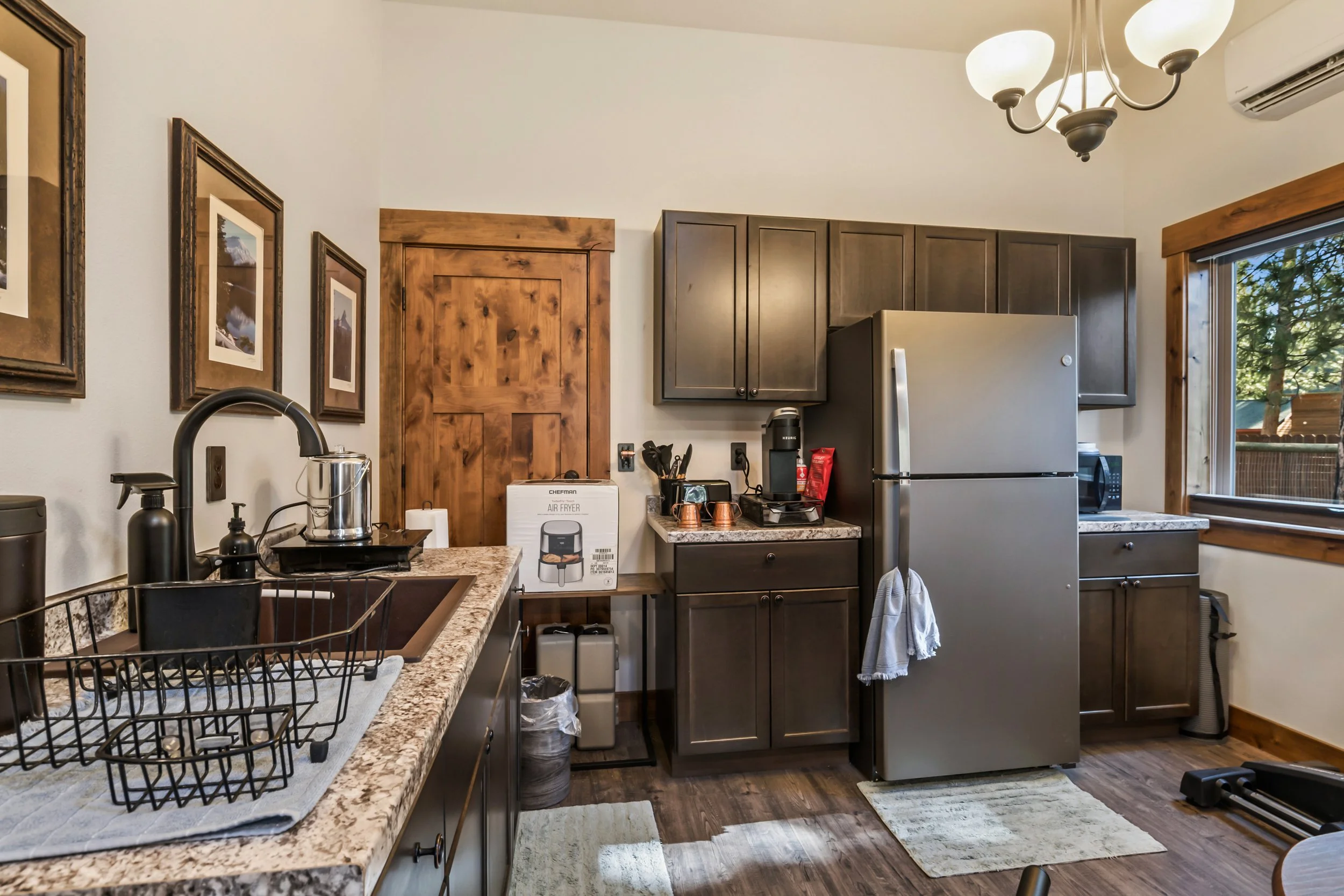 A kitchen with dark wooden cabinets, a granite countertop, a stainless steel refrigerator, a microwave, a coffee maker, and a window with wooden trim showing trees outside. There are framed pictures on the wall and a chandelier overhead.