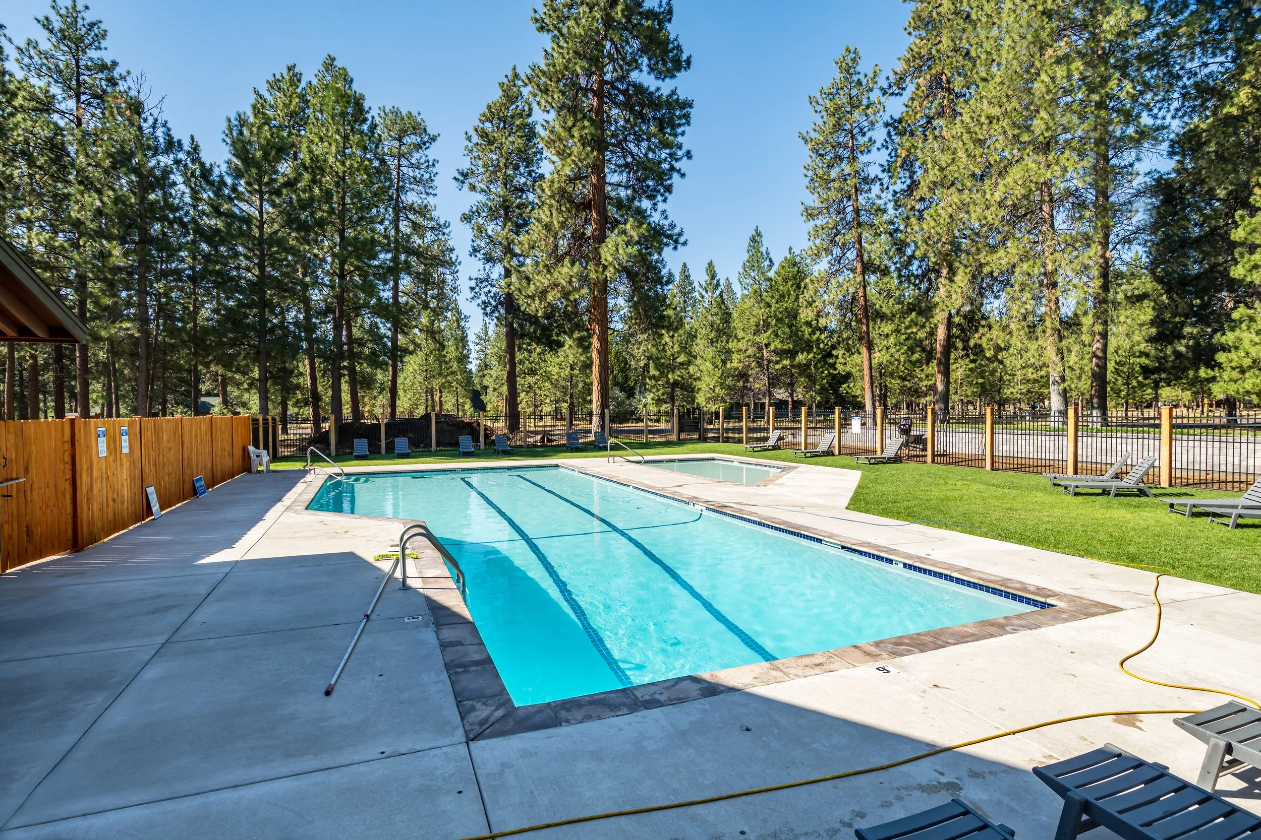 An outdoor swimming pool with a blue liner, surrounded by a concrete deck and lounge chairs, enclosed by a wooden fence, with tall pine trees and a clear blue sky in the background.