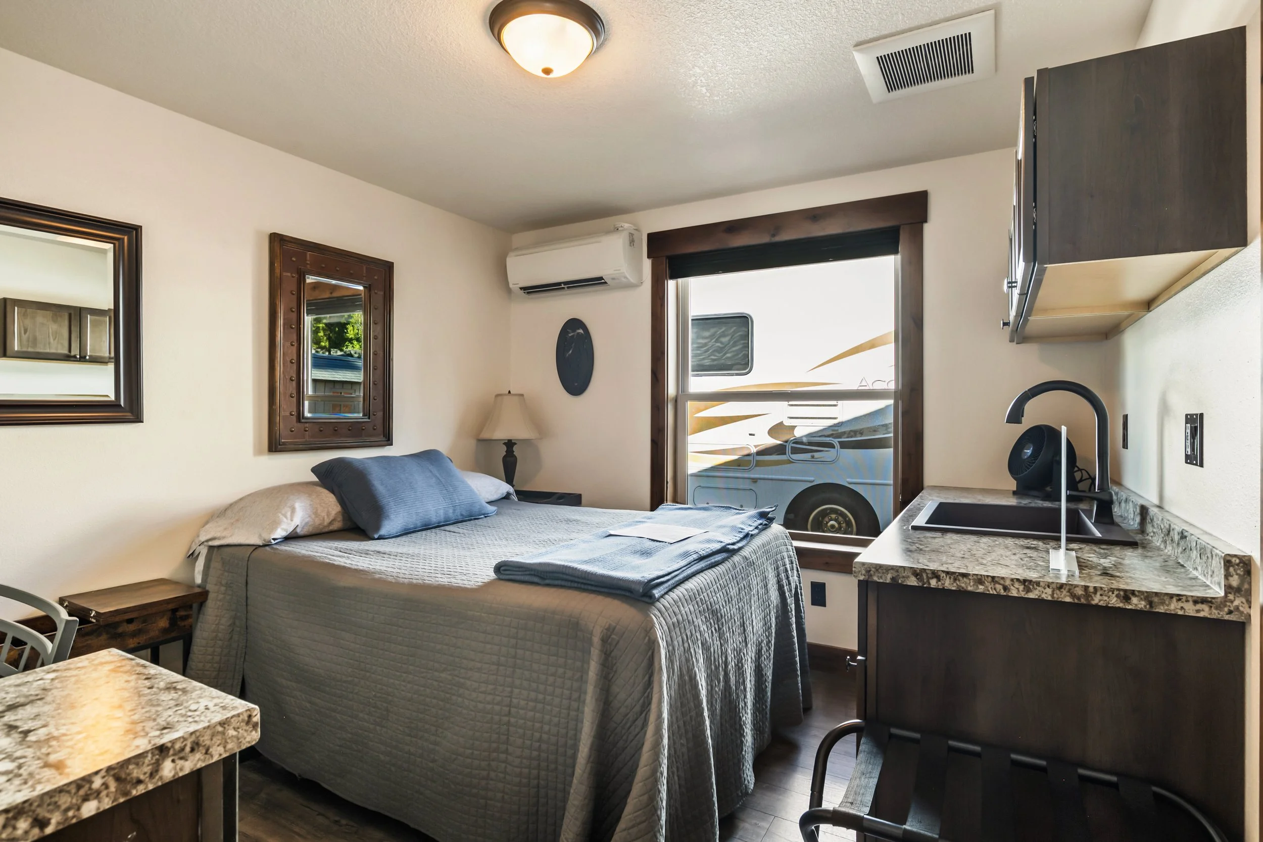 Interior of a bedroom with a bed, mirror, window, air conditioning unit, and kitchen counter with sink.