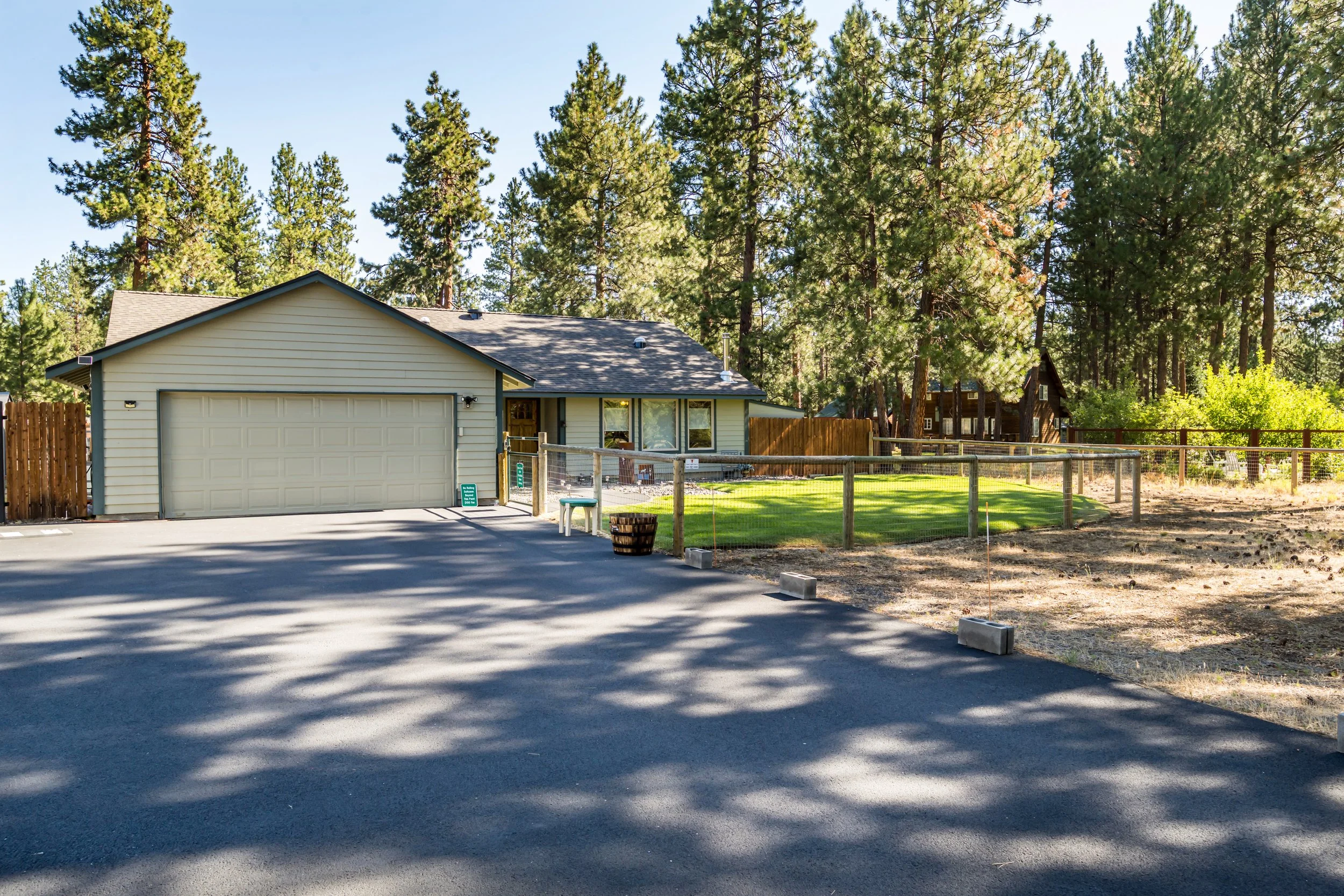 A residential house with a white garage door, surrounded by a wooden fence, tall pine trees, and a green lawn, under a clear blue sky.