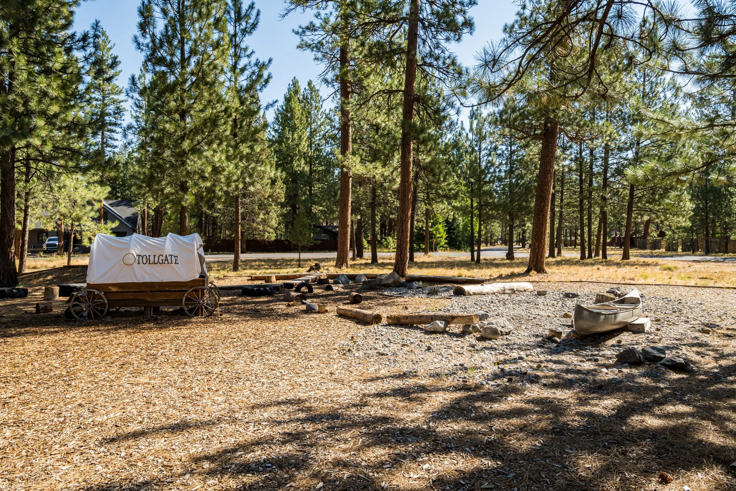 A forested area with tall pine trees and a gravel ground, a white tarp with 'TOLLGA' written on it covers an object, and a small silver rowboat rests on the ground.