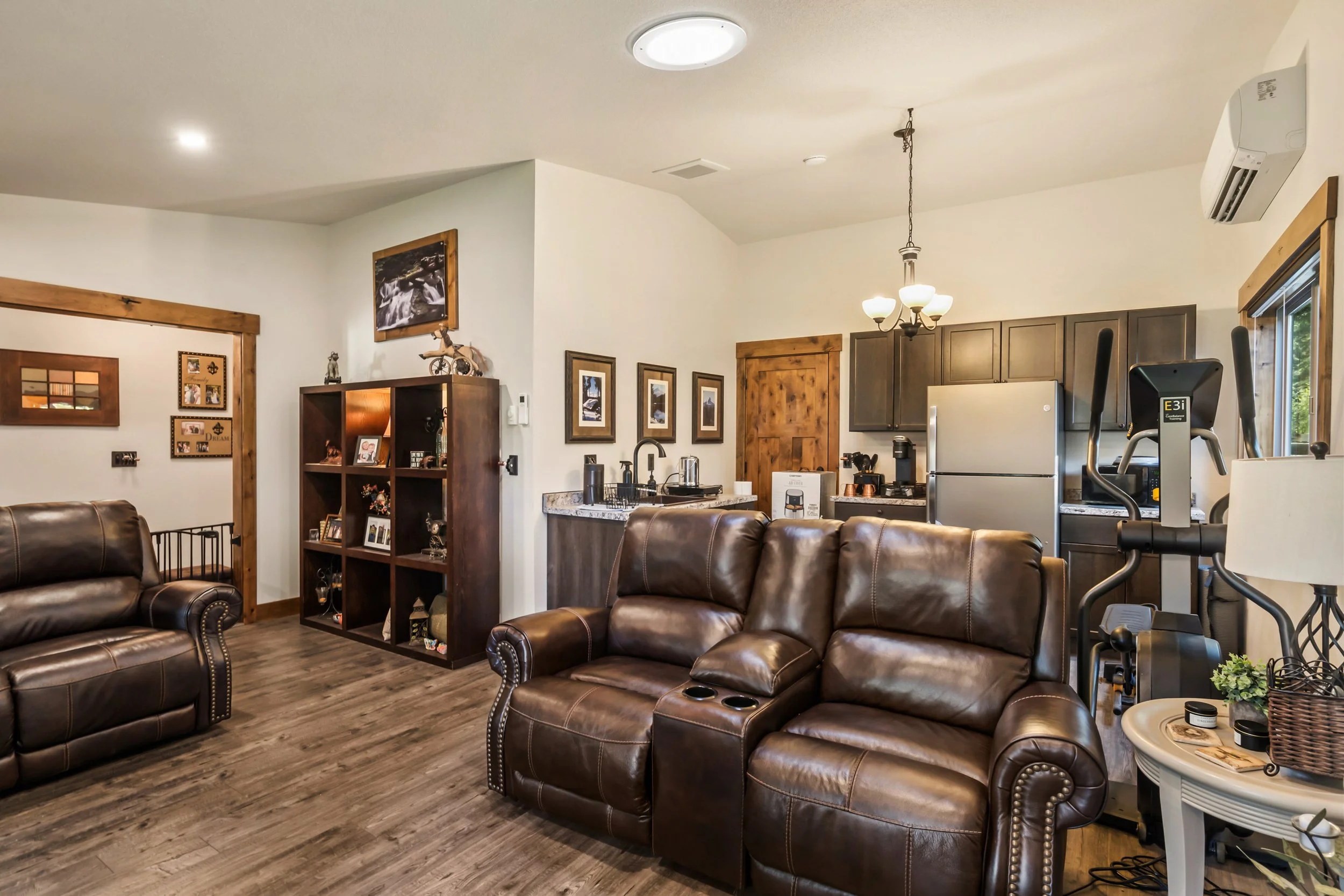 A cozy living room with brown leather sofas, a wooden floor, a wooden bookshelf, framed pictures on the walls, and a kitchen area with dark cabinets, a refrigerator, and various appliances.