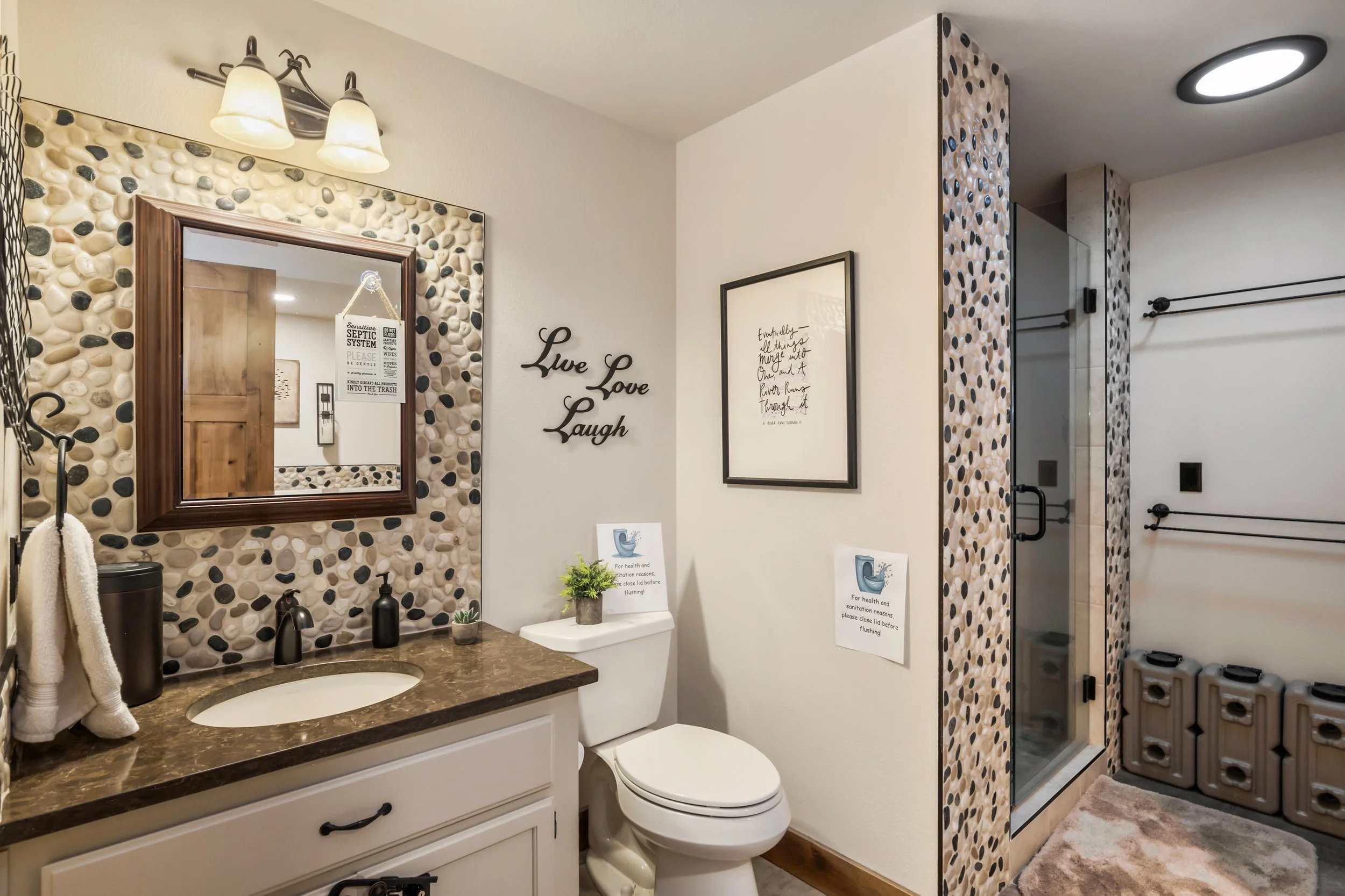 A bathroom with a pebble stone wall behind a mirror, a countertop with a sink and decor, a framed quote, and a toilet nearby. The shower area has a glass door, pebble wall, and a stack of washing machines in the corner.