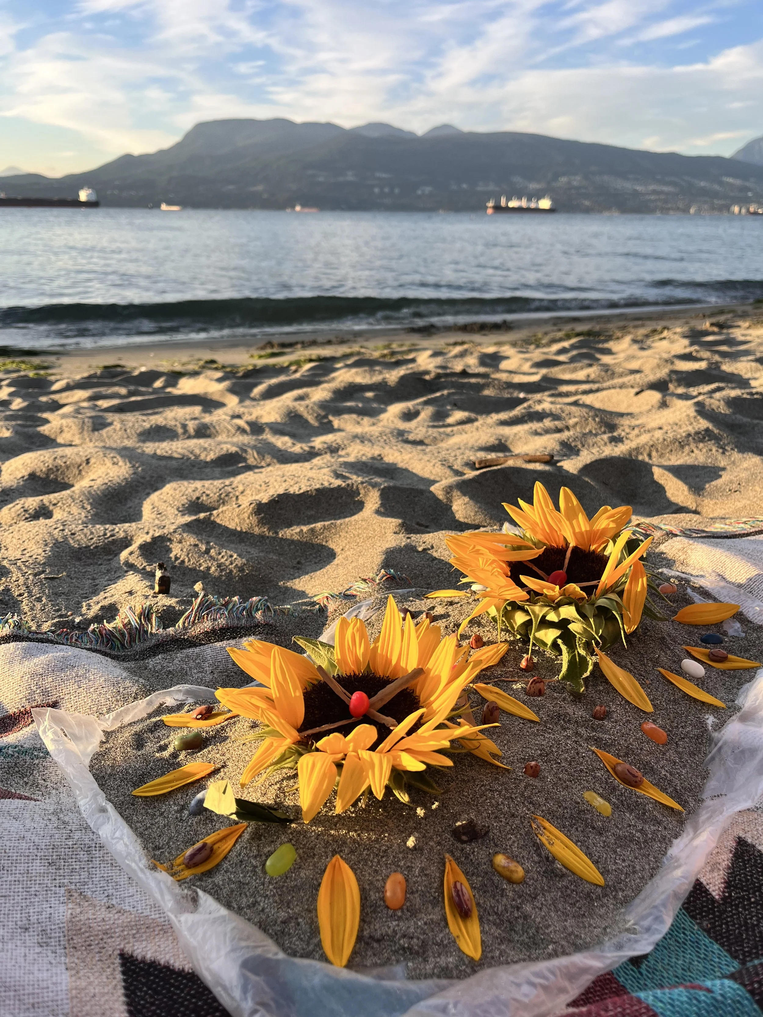 Colorful flower arrangement with yellow petals and red and green decorations on a sandy beach with a mountain and ships in the background.