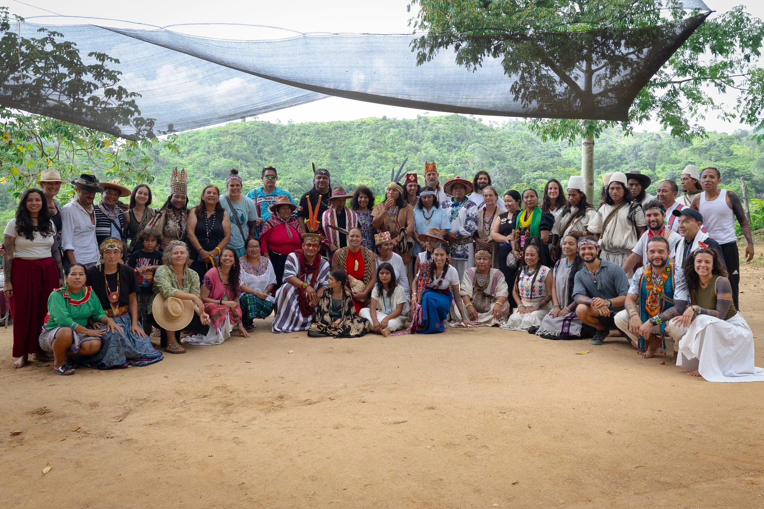 Group of Indigenous people and visitors dressed in traditional clothing and accessories, gathered outdoors under a large shade cloth with a lush green forested hillside in the background.