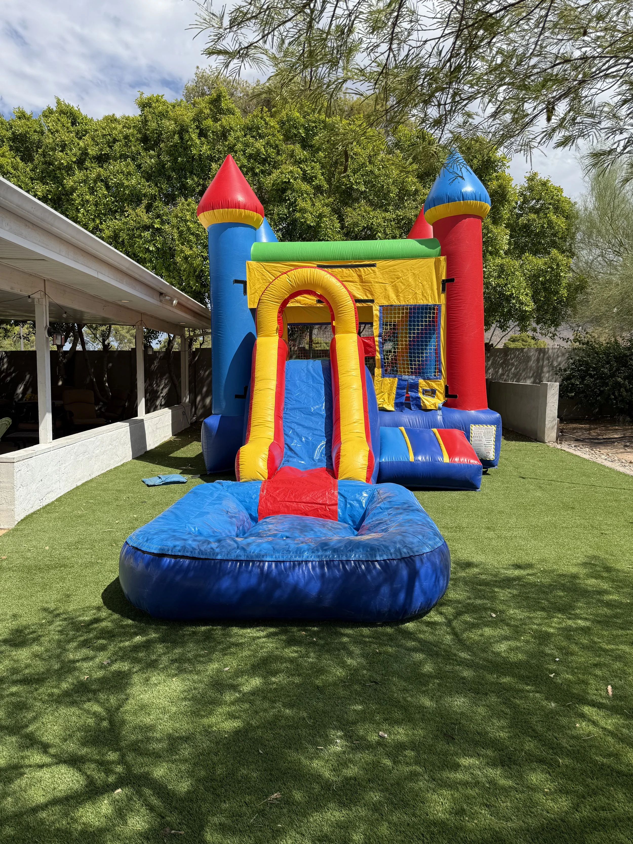 Colorful inflatable bounce house with slide set up on a grassy area outdoors, surrounded by trees.