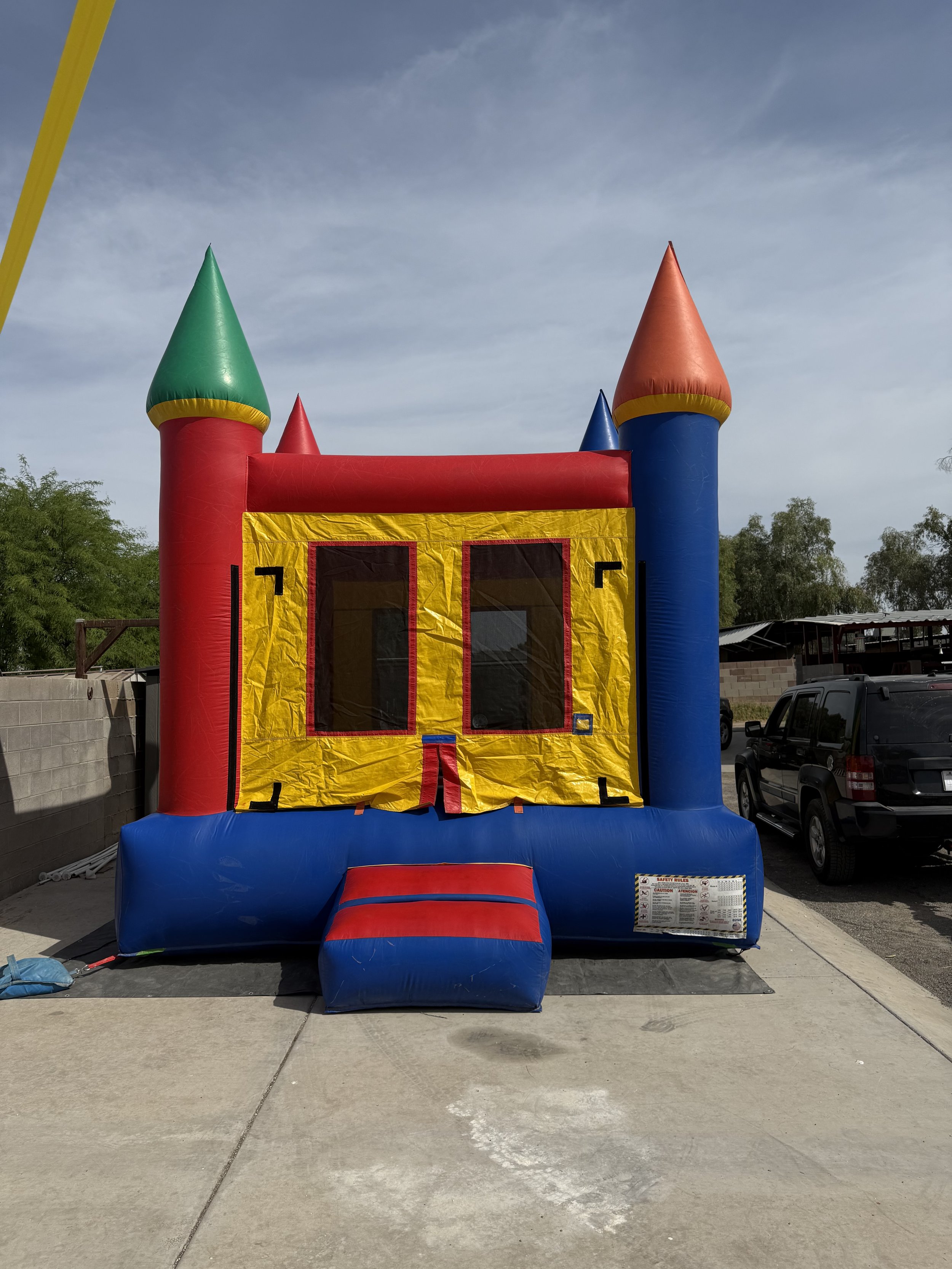 Colorful inflatable bounce house with castle turret design, yellow, red, green, blue, and orange colors, set on a concrete outdoor area with cars and trees in the background.