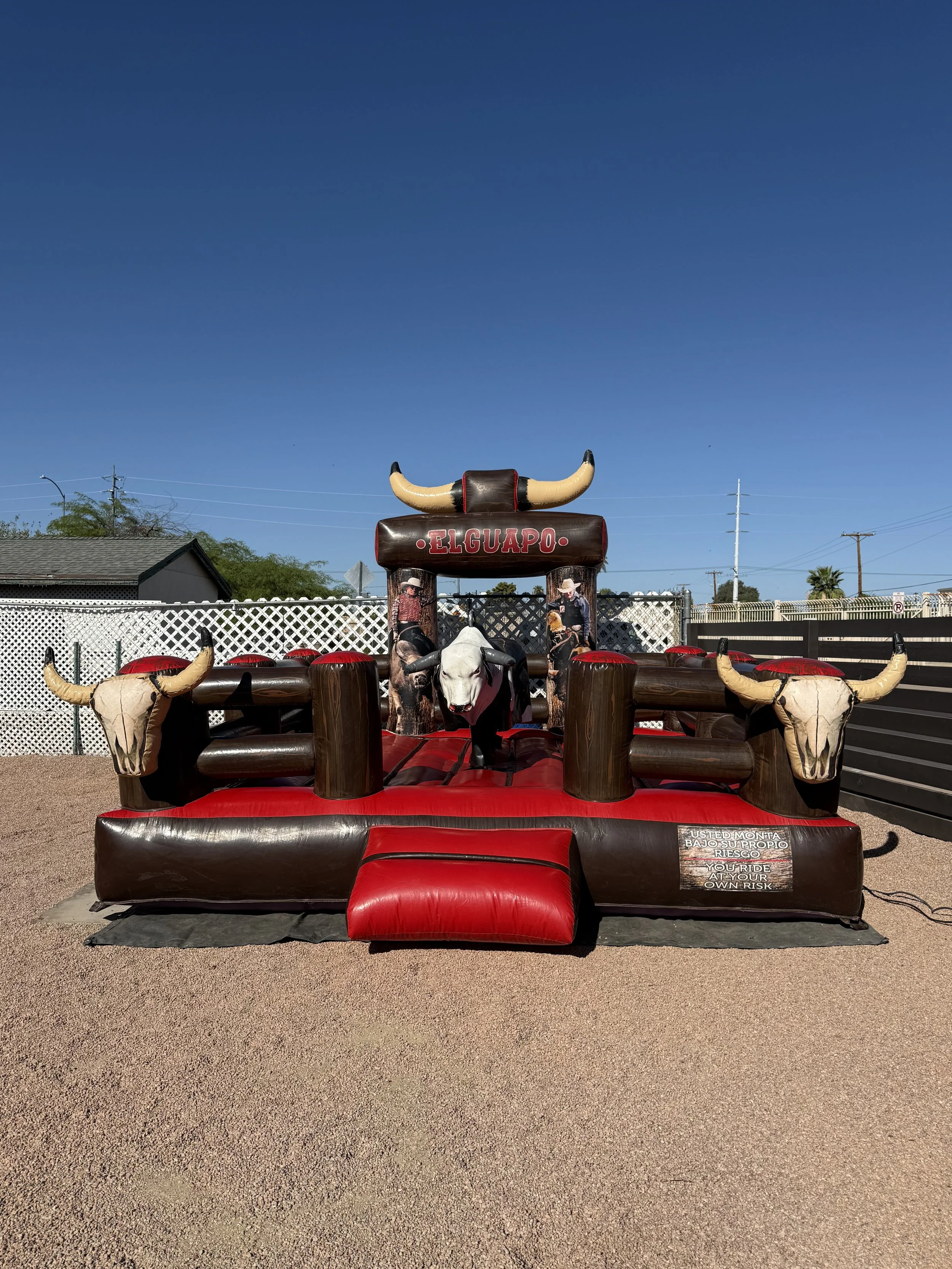 A large outdoor mechanical bull ride decorated with bull horns, skull images, and a sign that says "El Guapo." The ride has a red padded seat and black base, with a warning sign in Spanish and English that says "You ride at your own risk."