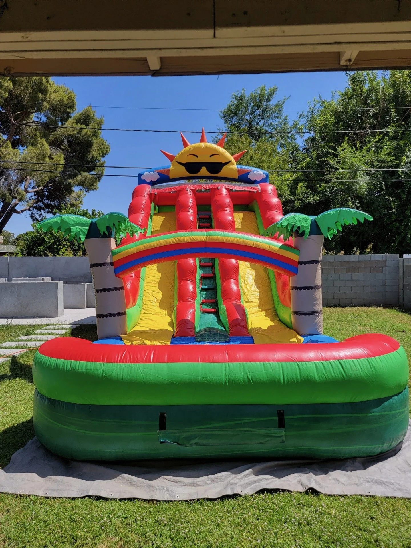 Colorful inflatable water slide with a smiling sun face wearing sunglasses at the top, set up on a lawn with trees and a brick wall in the background.