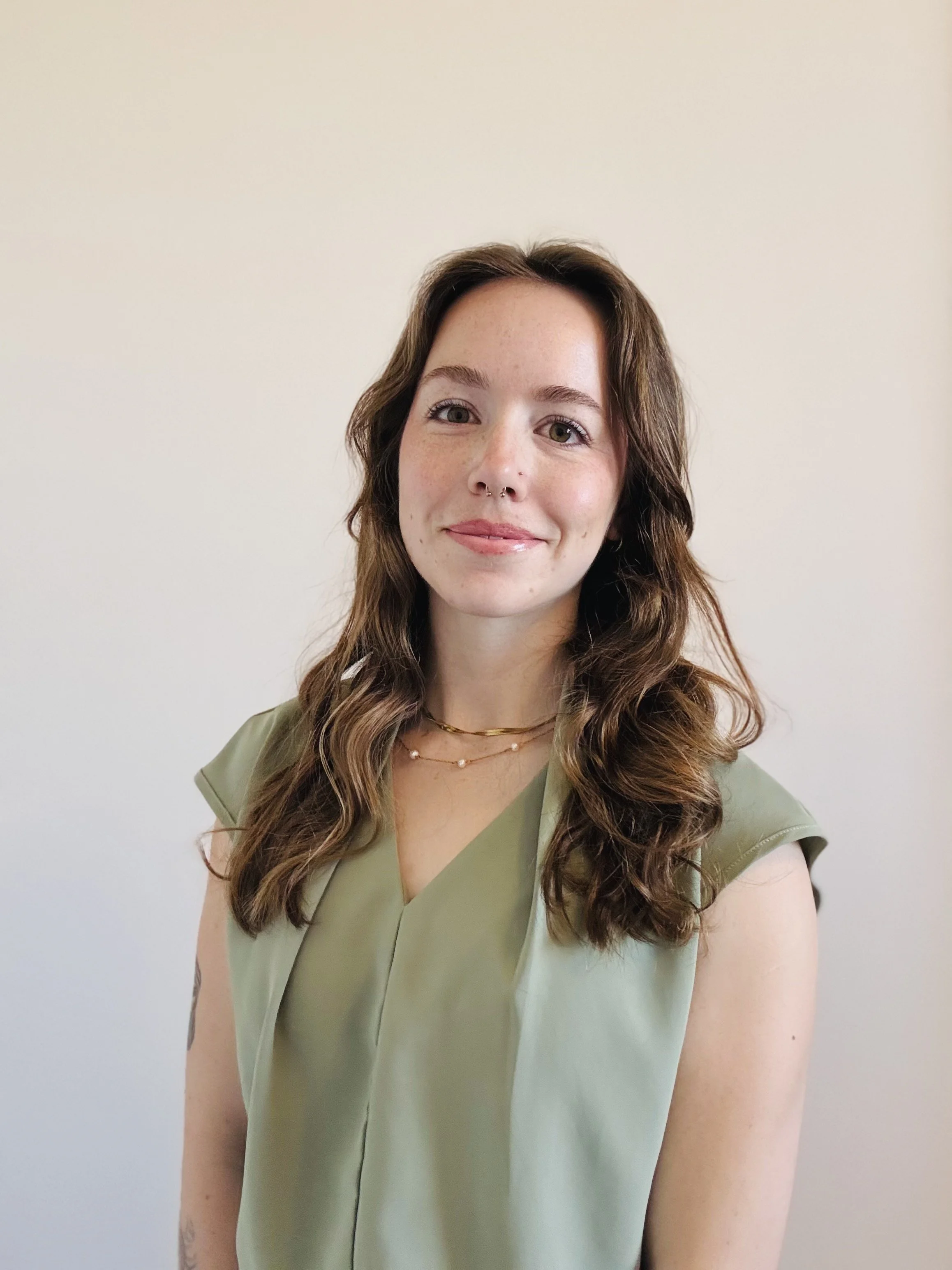 A picture of Allison: A young woman with wavy brown hair, light skin, freckles, and a septum piercing, smiling slightly, wearing a light green sleeveless top and layered necklaces, standing against a plain white wall.