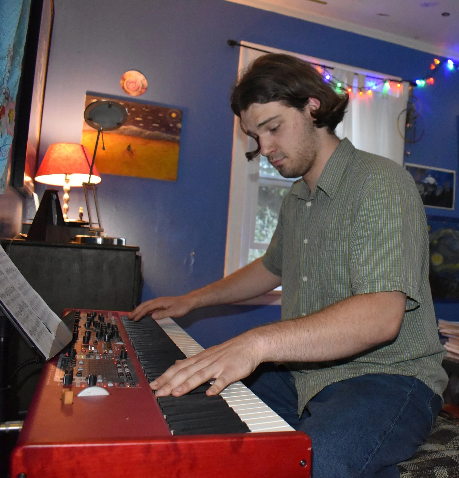 Young man playing a keyboard in a dimly lit room with blue walls, a red lampshade on a small table, and colorful string lights. Artwork is hung on the walls.
