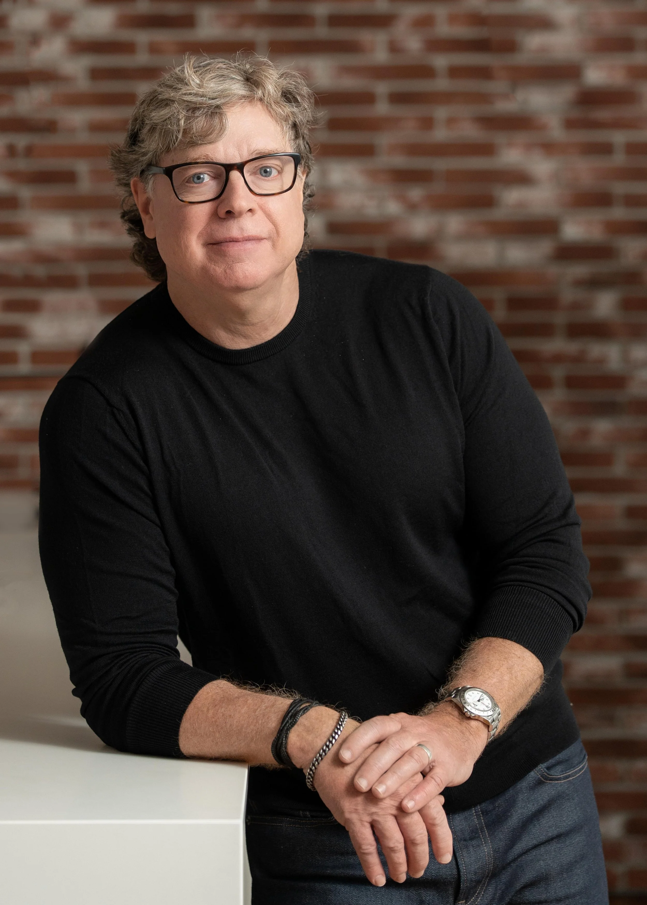 A middle-aged man with gray curly hair wearing glasses, a black long-sleeve shirt, and jeans, leaning on a white surface with a brick wall in the background.