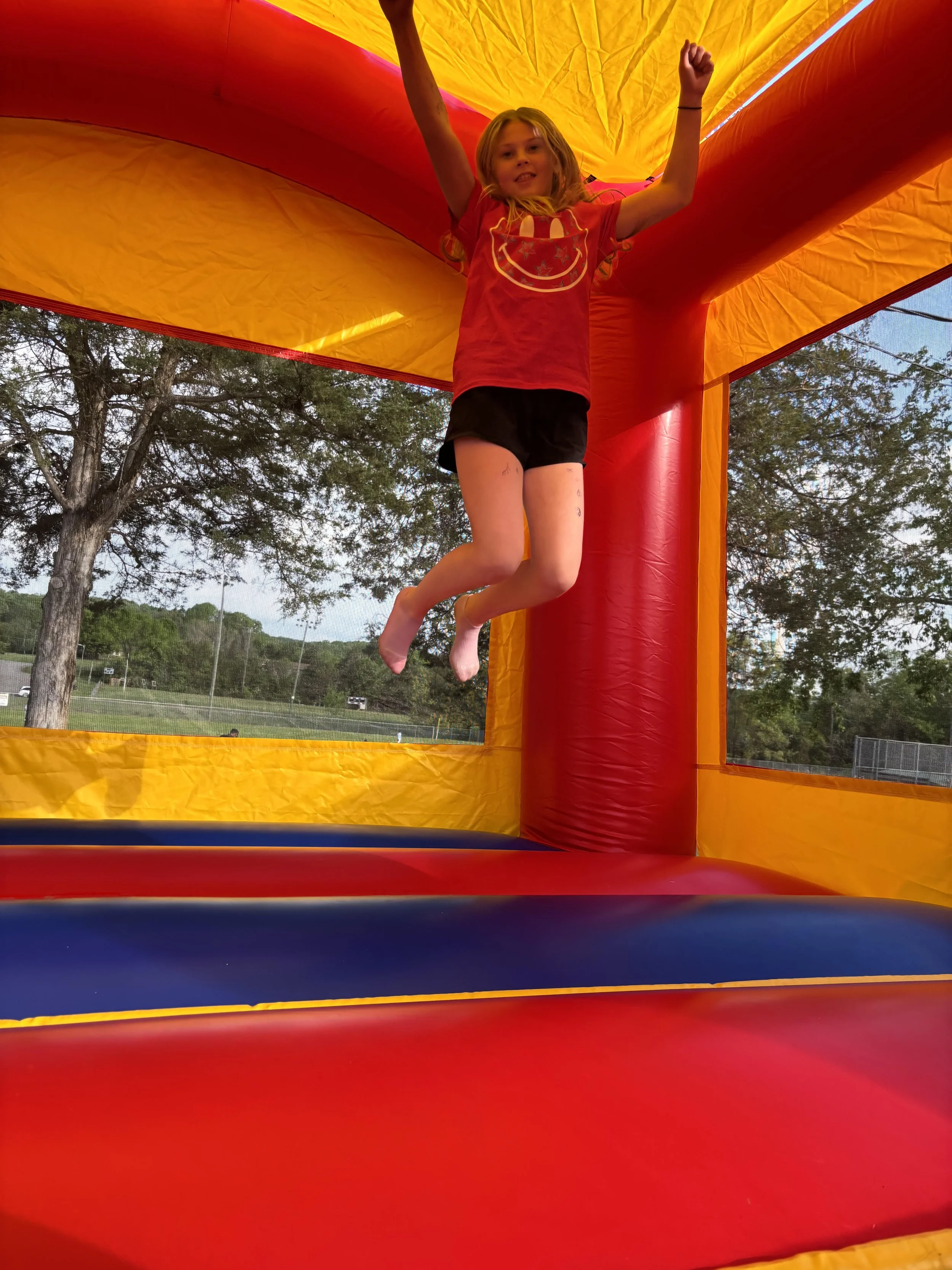 A young girl with blonde hair wearing a red shirt with a smiley face, hanging from the ceiling inside an orange and yellow inflatable bounce house. Outside, trees and a park are visible.