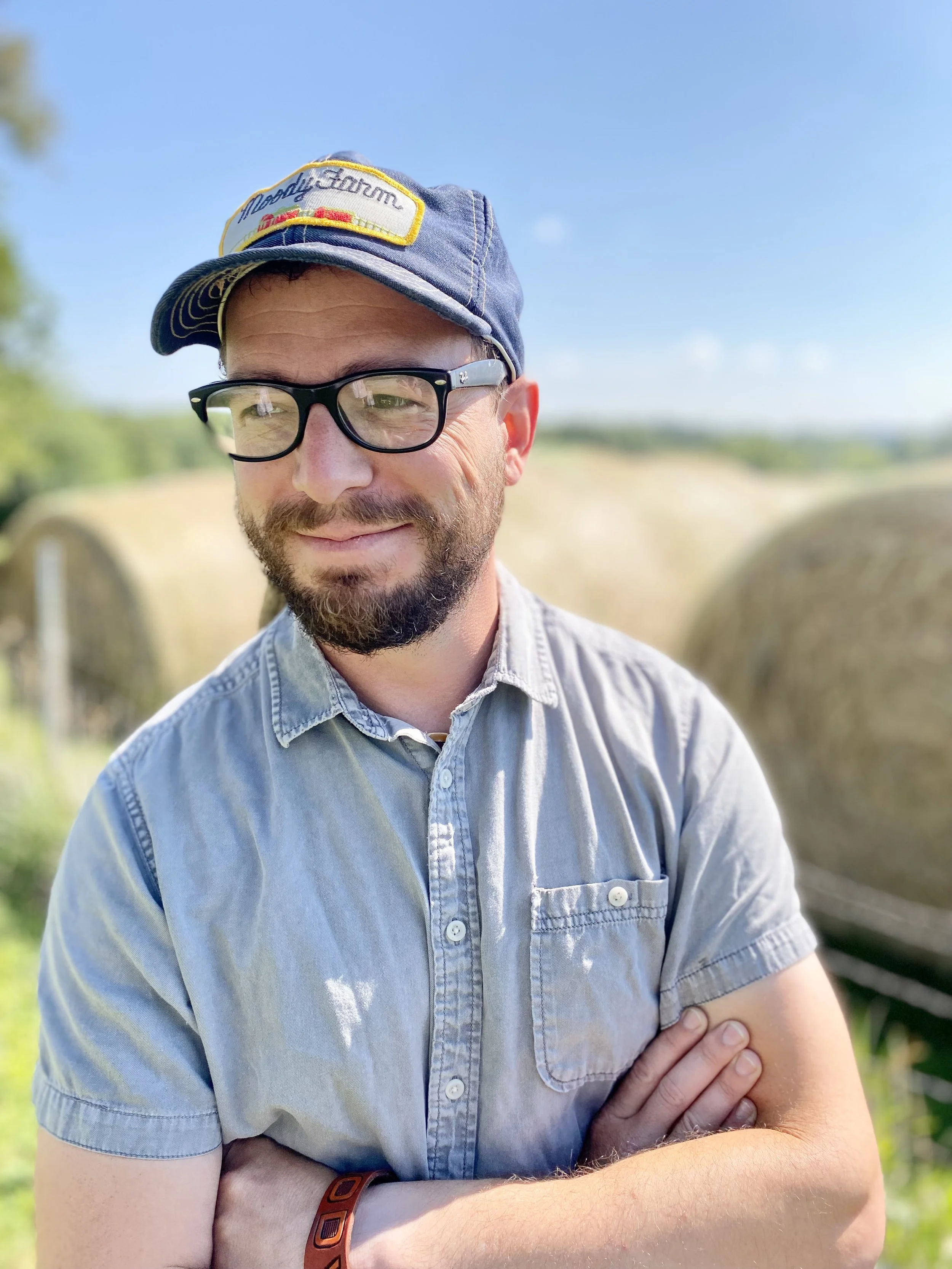 A man with glasses and a beard wearing a denim cap and a light blue denim shirt standing outdoors with greenery and blue sky in the background.