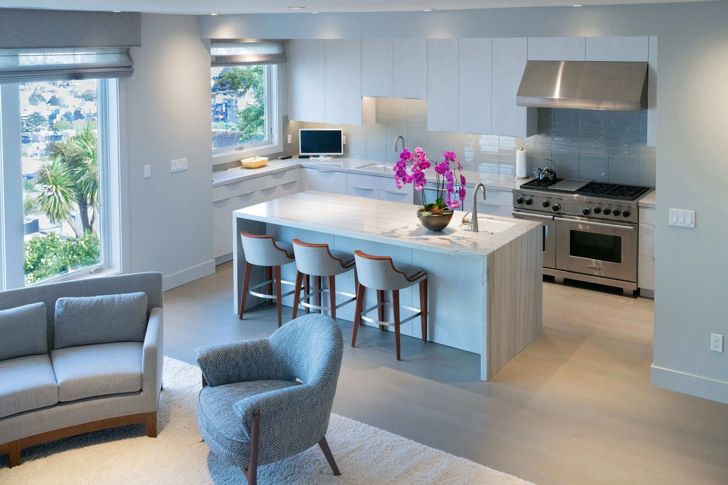 Bright kitchen with white cabinets, stainless steel appliances, a central island with a pink orchid, and a view of a neighborhood through large windows.