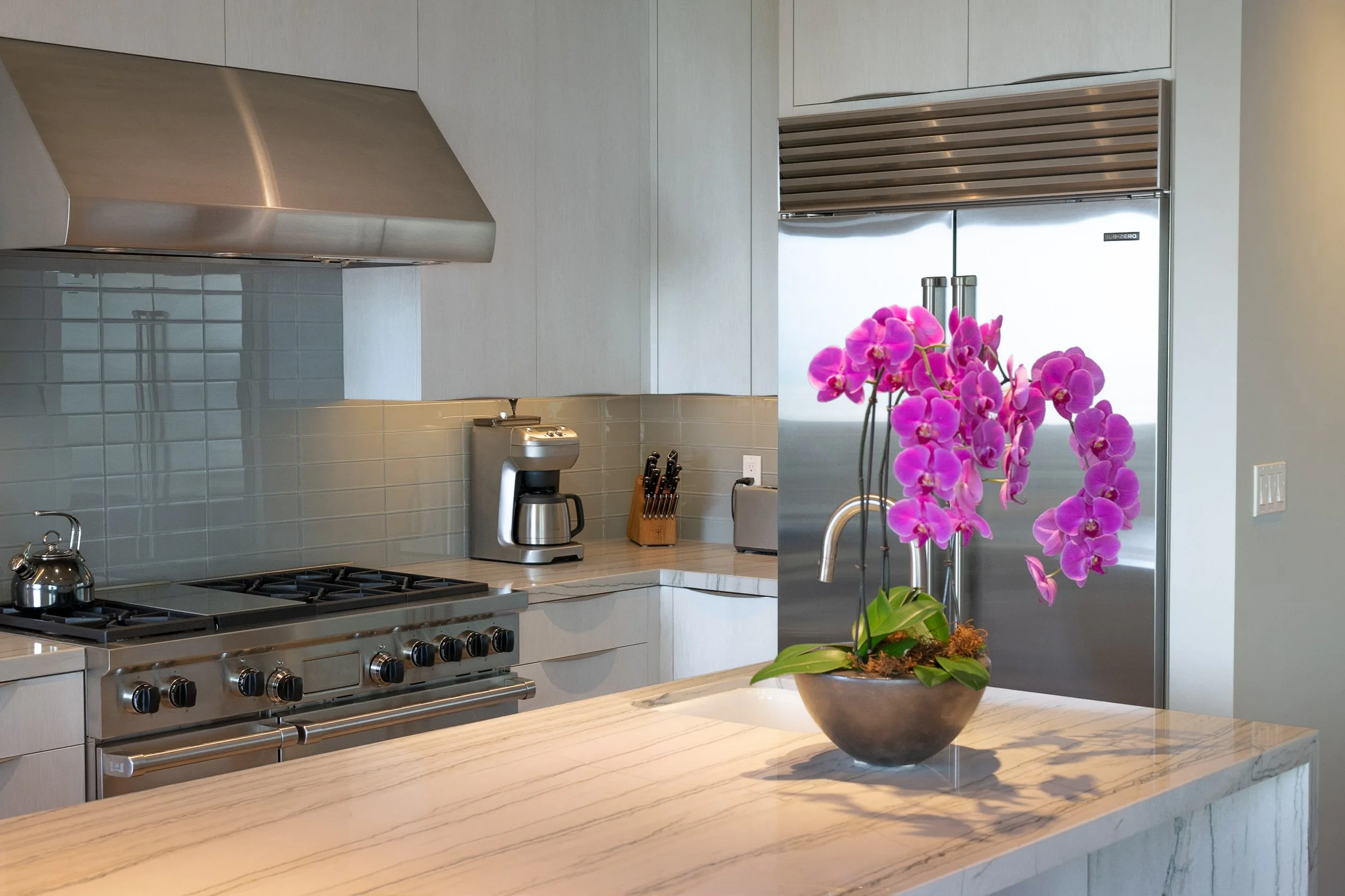 A modern kitchen with light gray cabinets, stainless steel appliances, and a marble countertop. There is a purple orchid plant in a gray pot on the countertop, a coffee maker, a knife block, and a stainless steel refrigerator. The backsplash is made 