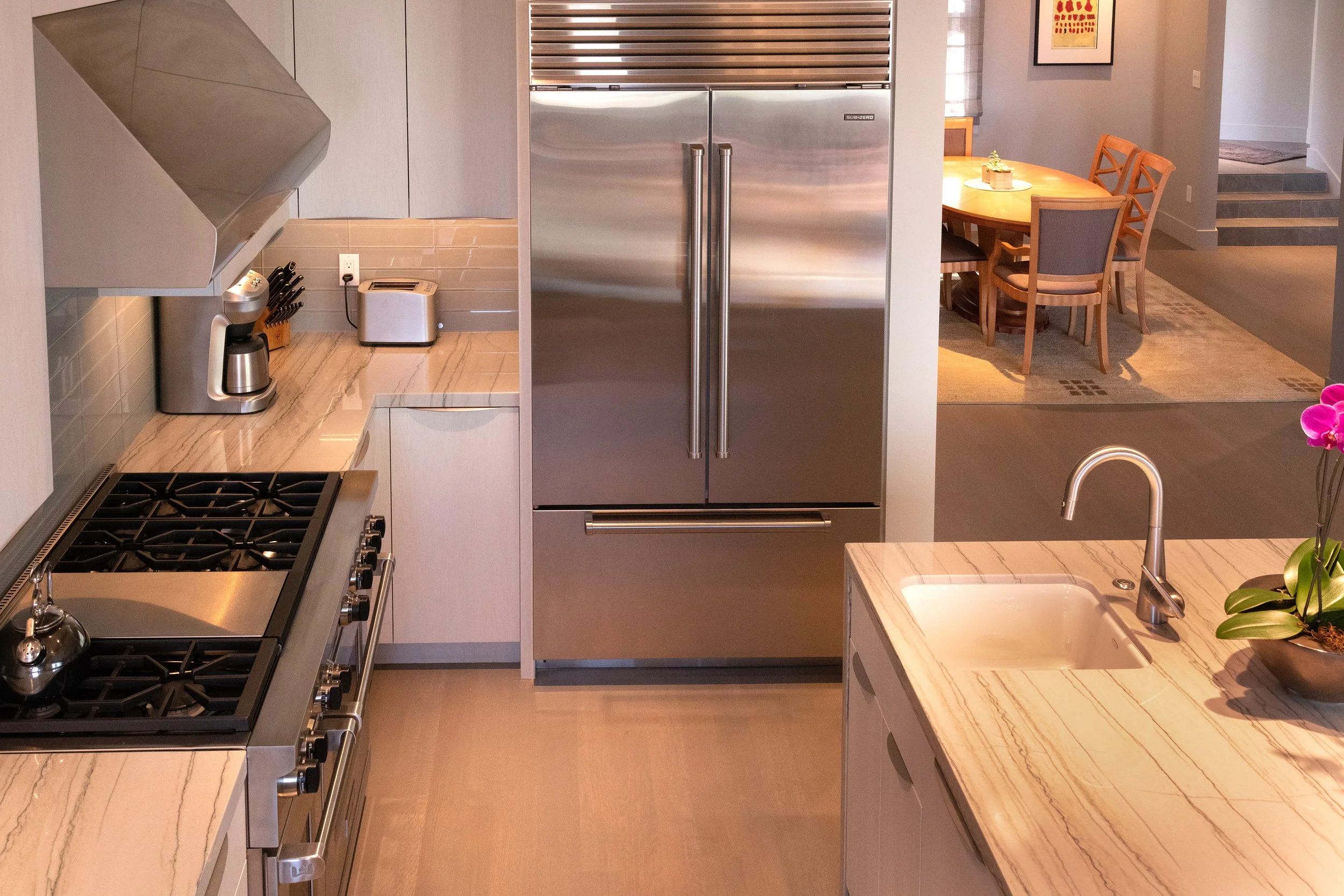 Modern kitchen with a gas stove, a marble countertop, a stainless steel refrigerator, a sink with a faucet, and a dining area with a wood table and chairs. There is a potted orchid on the counter and a framed artwork on the wall.