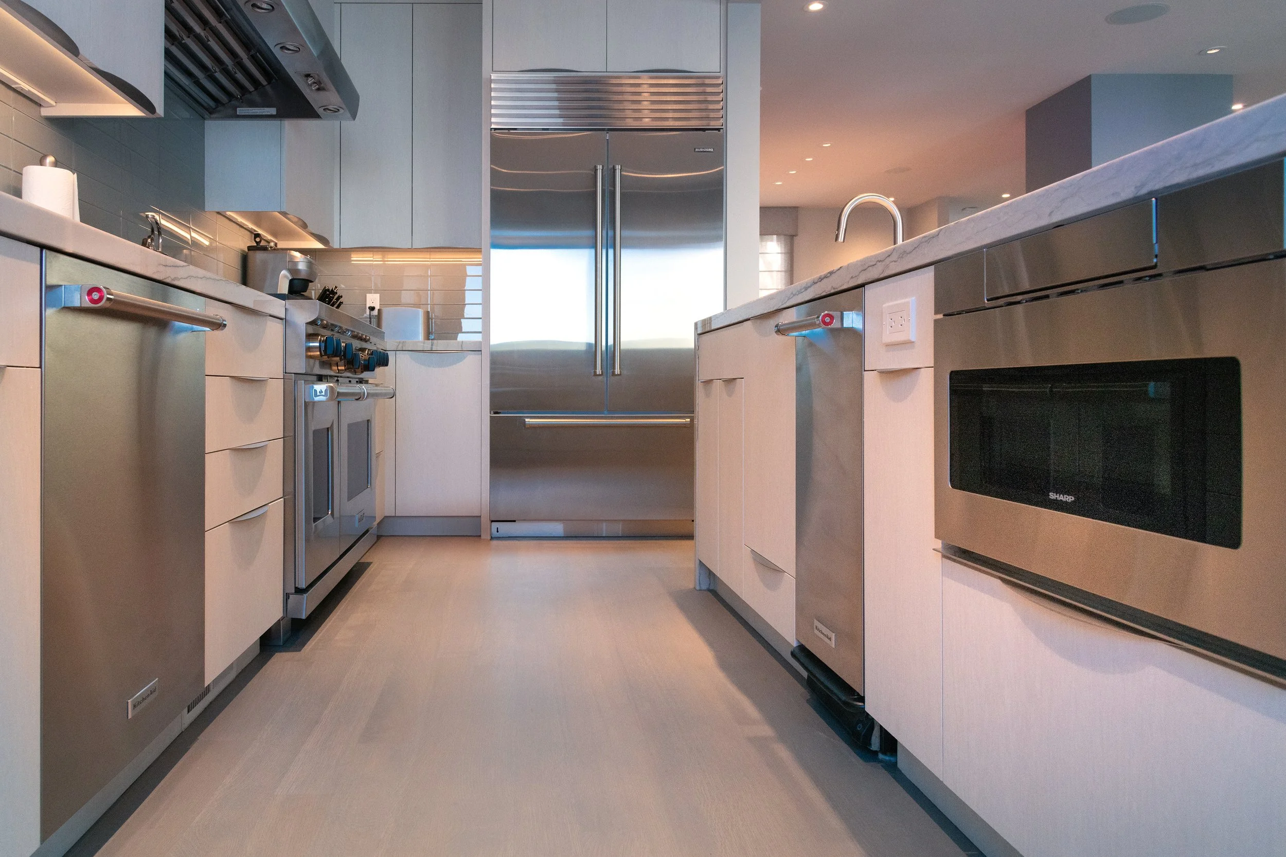 Modern kitchen with stainless steel appliances, light-colored cabinetry, marble countertops, and beige flooring.