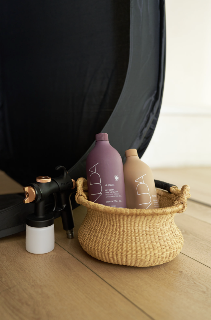 Two bottles of Aveda hair care products in a woven basket, with a photography lighting reflector and spray gun nearby on a wooden floor.