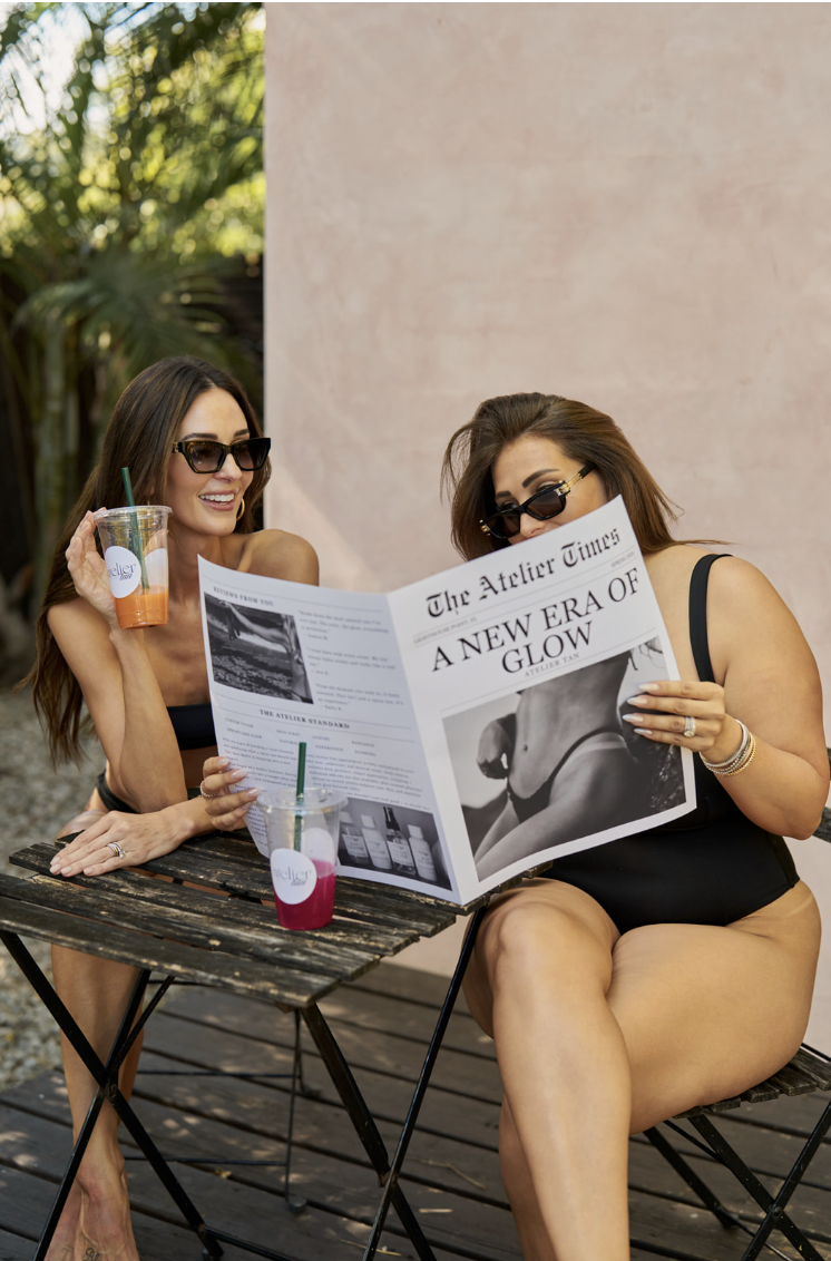 Two women sitting at a wooden table outdoors, wearing swimsuits and sunglasses, reading a newspaper titled 'The Atelier Times' with the headline 'A NEW ERA OF GLOW', each holding a drink with straws.
