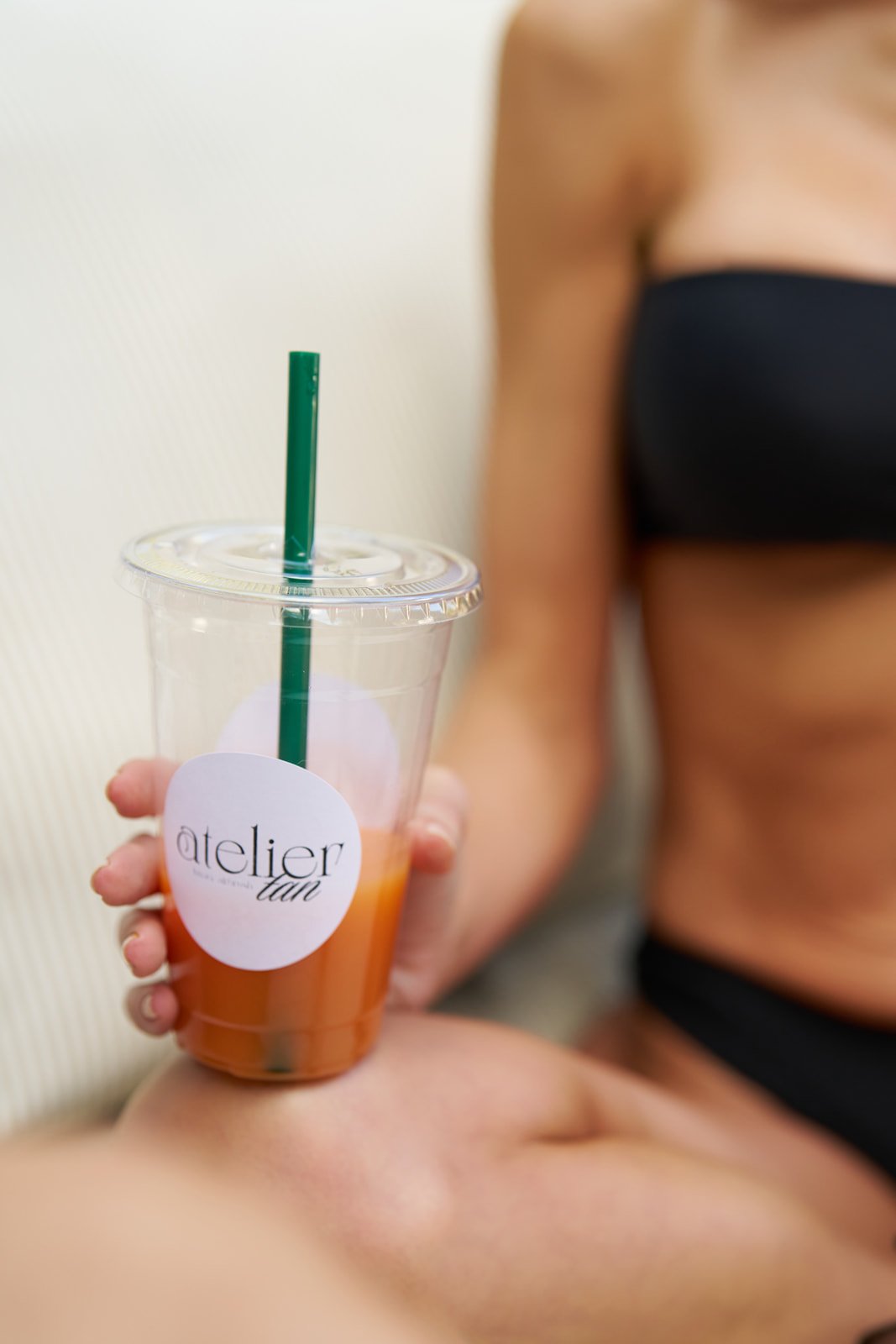 Person holding a plastic cup with orange beverage and green straw, wearing black clothing, with part of their arm and torso visible.