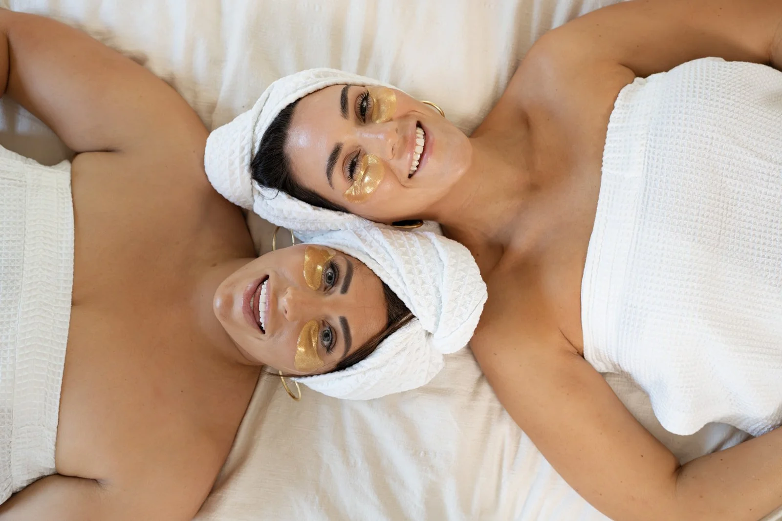Two women are lying on a bed with white towels wrapped around their heads and white spa robes. They are smiling and have gold eye masks on their faces.