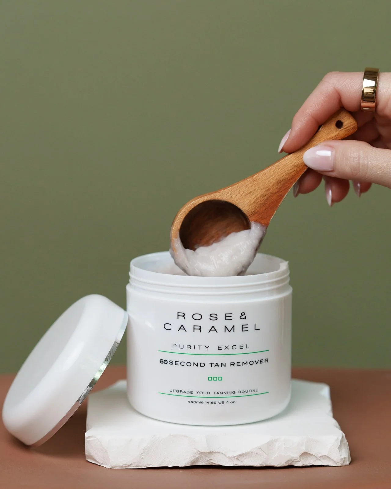 A hand with a gold ring is scooping a white cream from a container labeled "Rose & Caramel Purity Excel 60 Second Tan Remover." The container is placed on a white block with a green background visible.