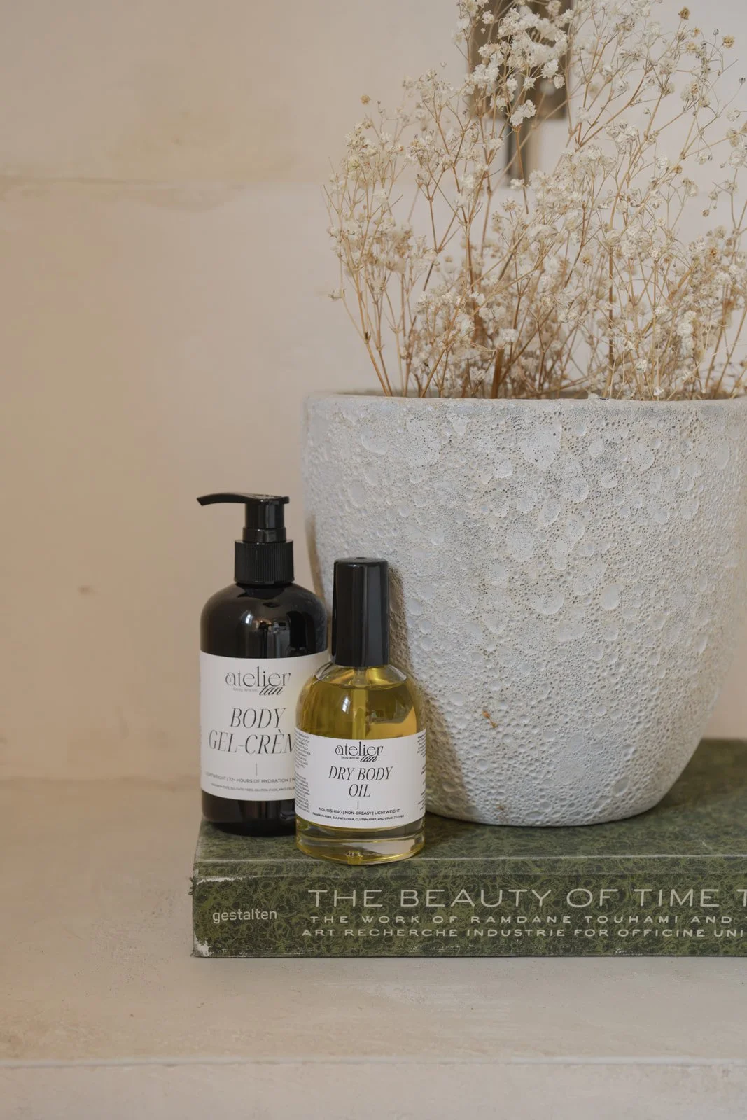 A large textured white pot with dried white flowers, two black and white labeled skincare bottles (body gel cream and dry body oil) are placed in front of the pot, and a green book titled 'The Beauty of Time' is beneath them.