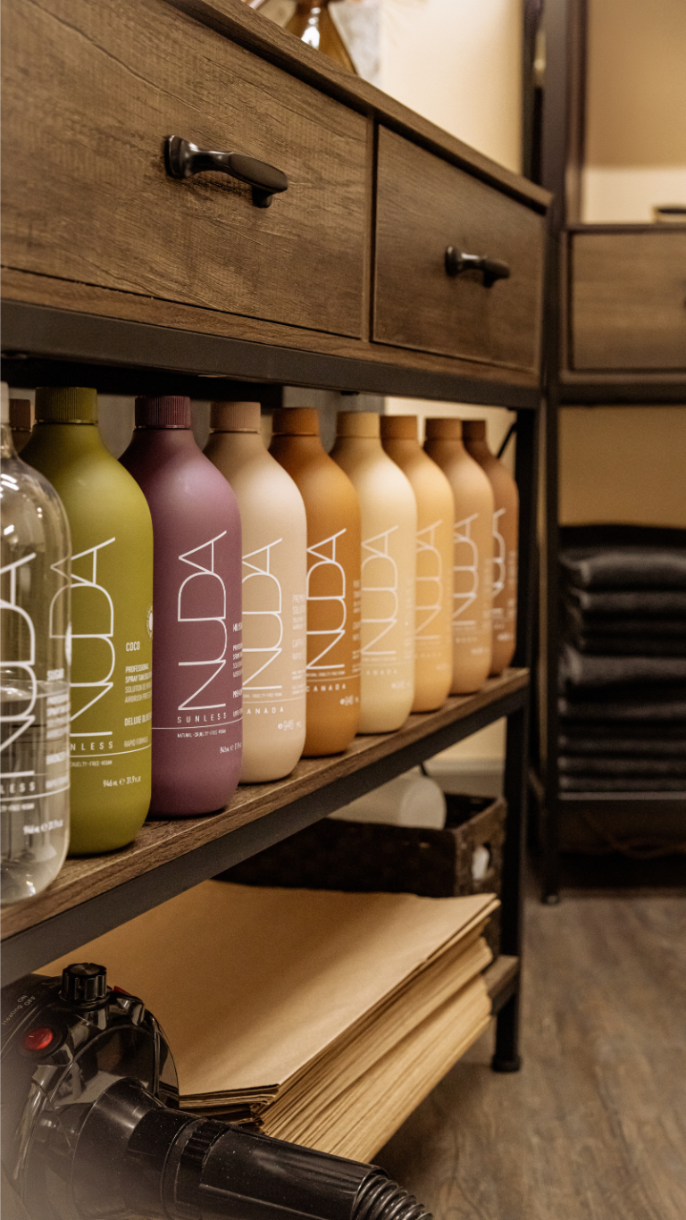 Row of colorful hair care bottles on a wooden shelf in a salon, with a hair dryer and black towels in the background.