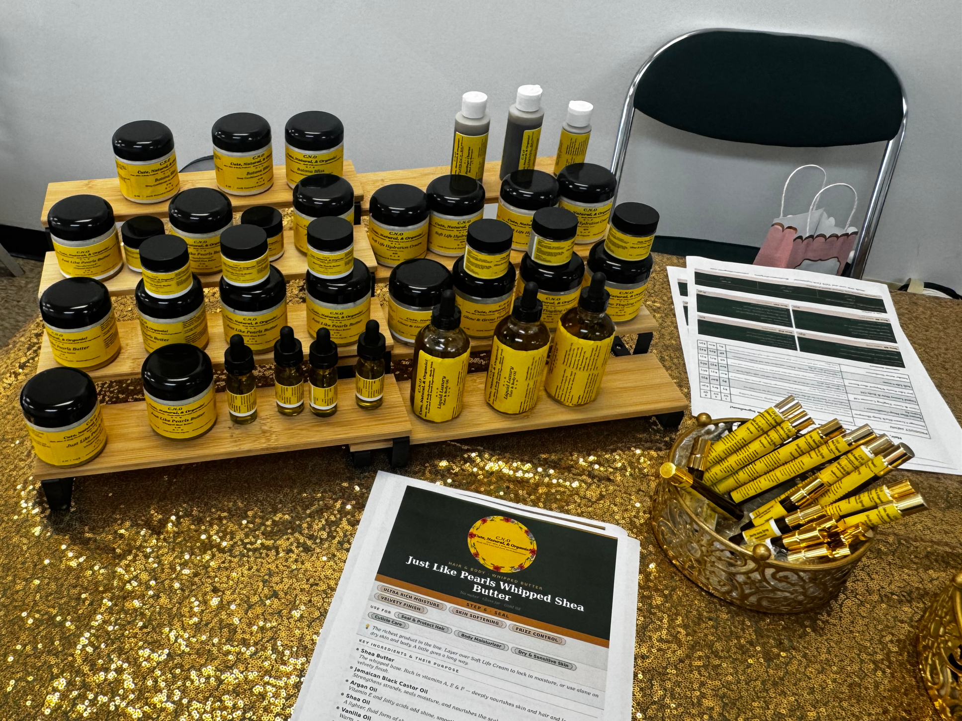 Display of small jars and bottles of shea butter with yellow labels, arranged on wooden stands, next to a brochure titled "Just Like Pearls Whipped Shea Butter" on a gold sequin tablecloth.