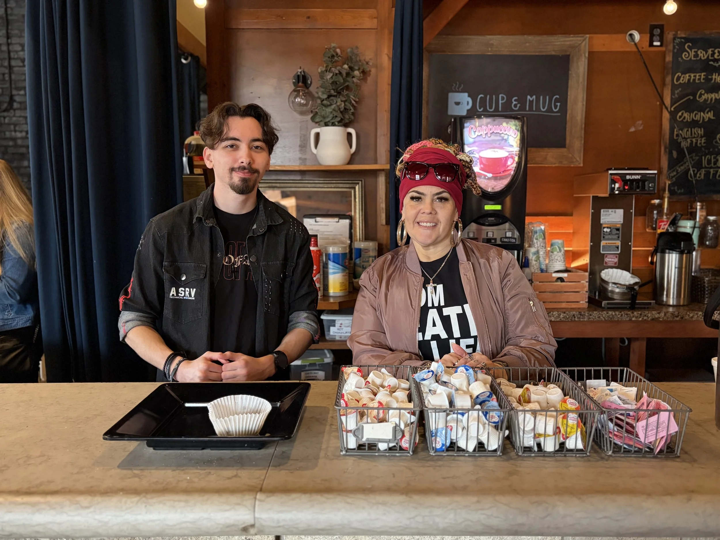 Two people stand behind a counter with baskets of coffee creamer cups. The background features a coffee shop interior with wooden walls, a sign reading 'CUP & MUG,' and a chalkboard menu.