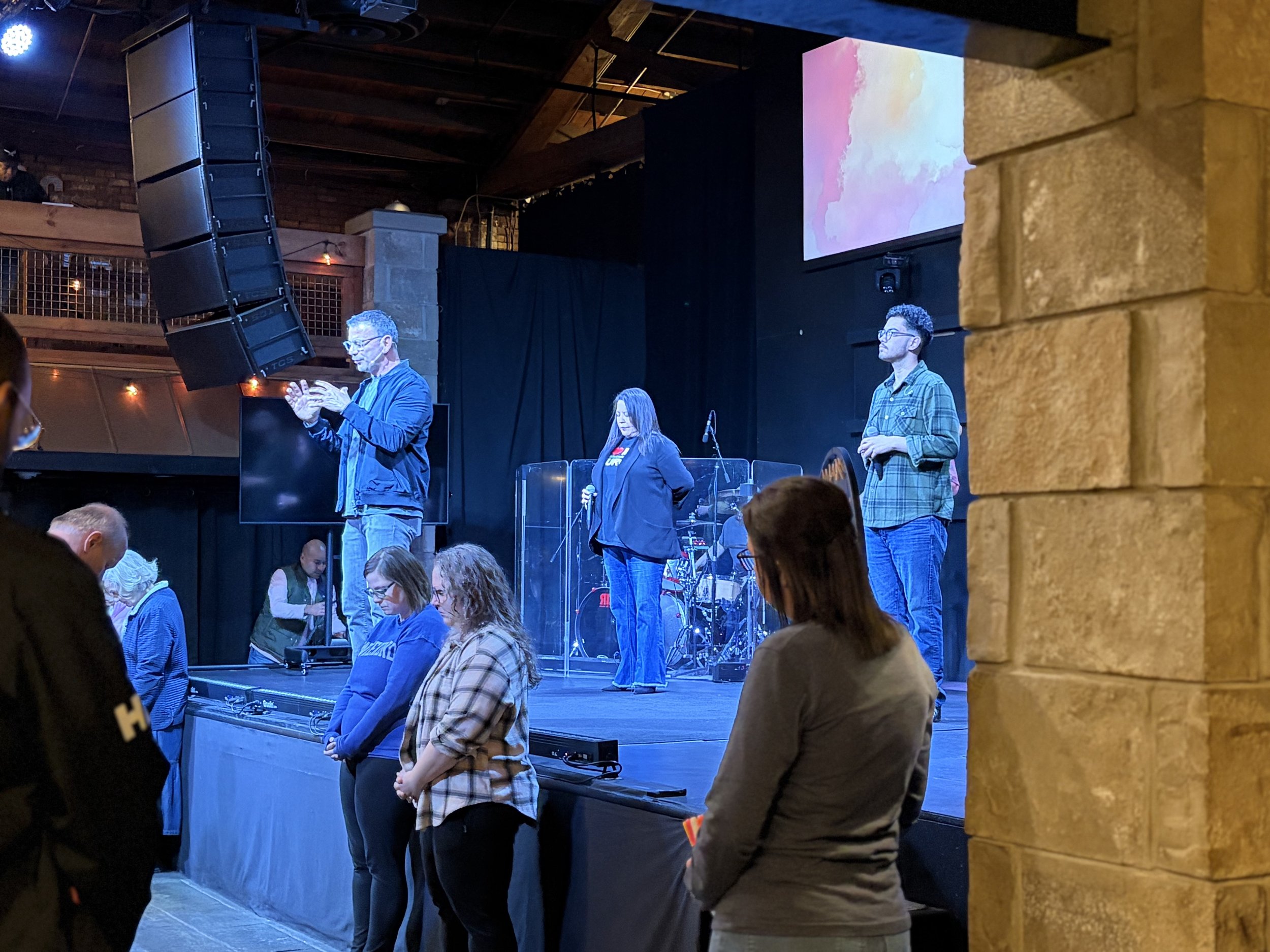 People standing indoors during a presentation or event, with a large screen displaying abstract pastel colors. The image shows attendees with some on the stage, and a speaker or presenter at a microphone behind a clear shield.