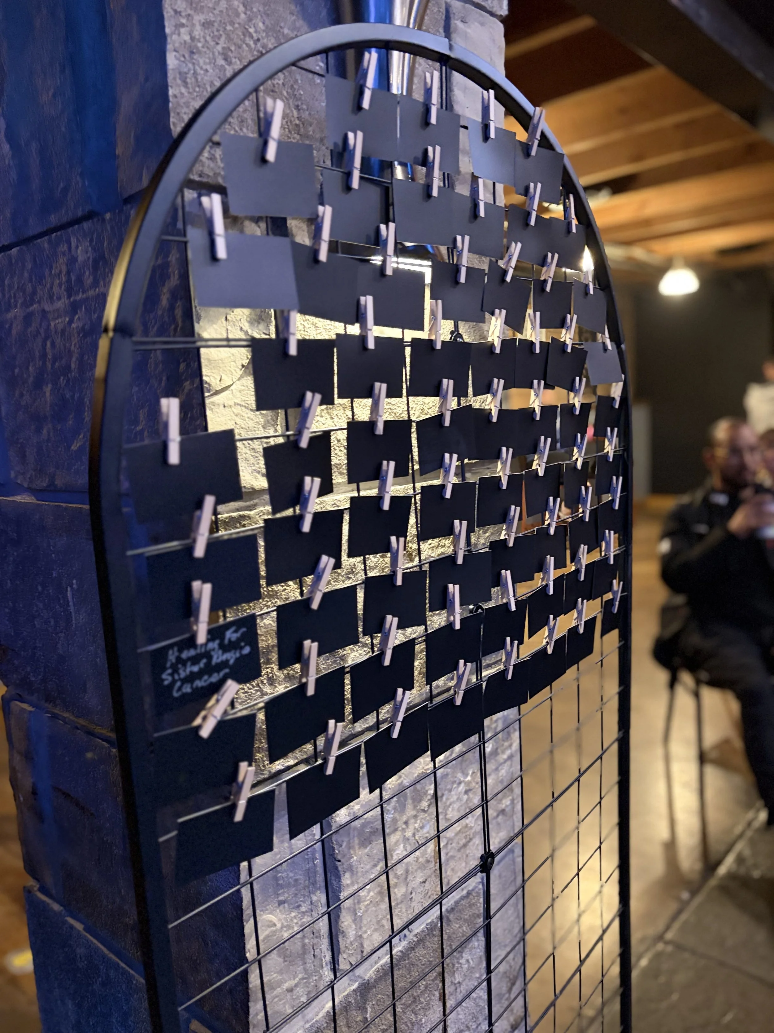 Black photo holder board with small clothespins, attached to a metal grid, mounted on a stone wall. The background shows a dimly lit room with wooden ceiling beams and a few people in the distance.