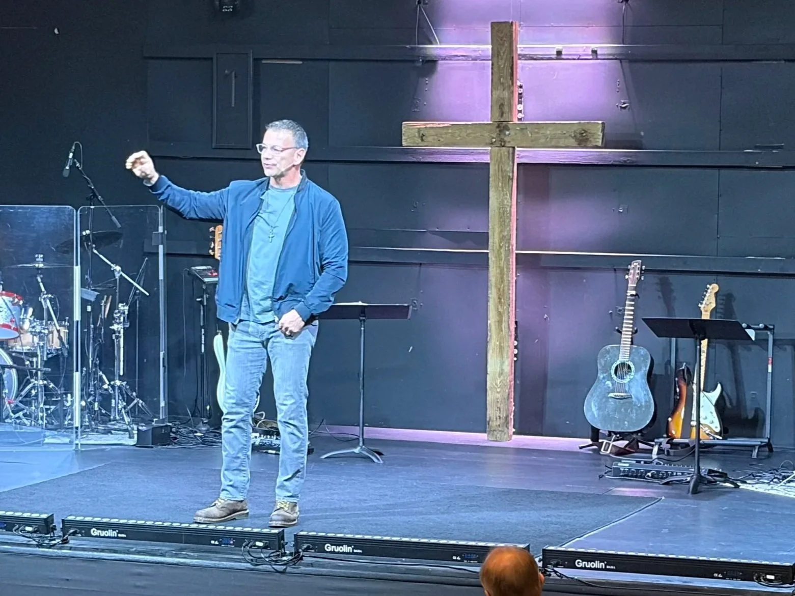 A man standing on a stage with musical instruments, a large wooden cross in the background, and purple lighting.