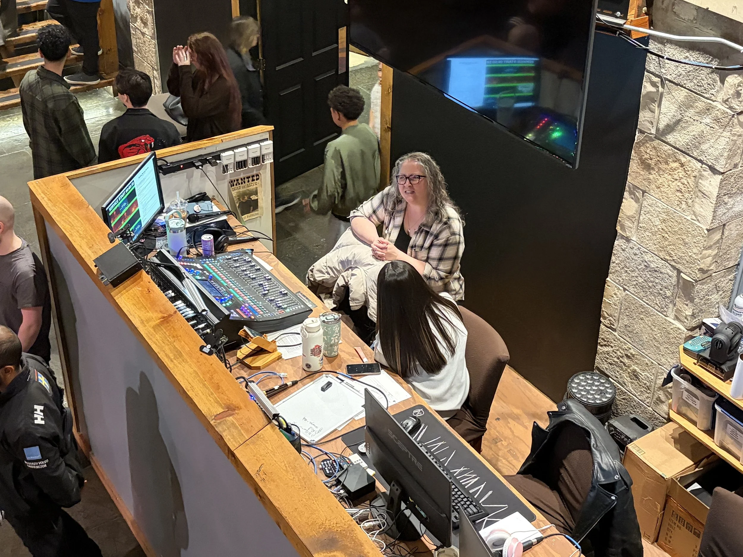 A woman with glasses and curly hair sitting at a desk with computer equipment, a notepad, and drinks on it, in a room with a stone wall. Several people are standing and talking behind her, with some near a black wall and monitor.