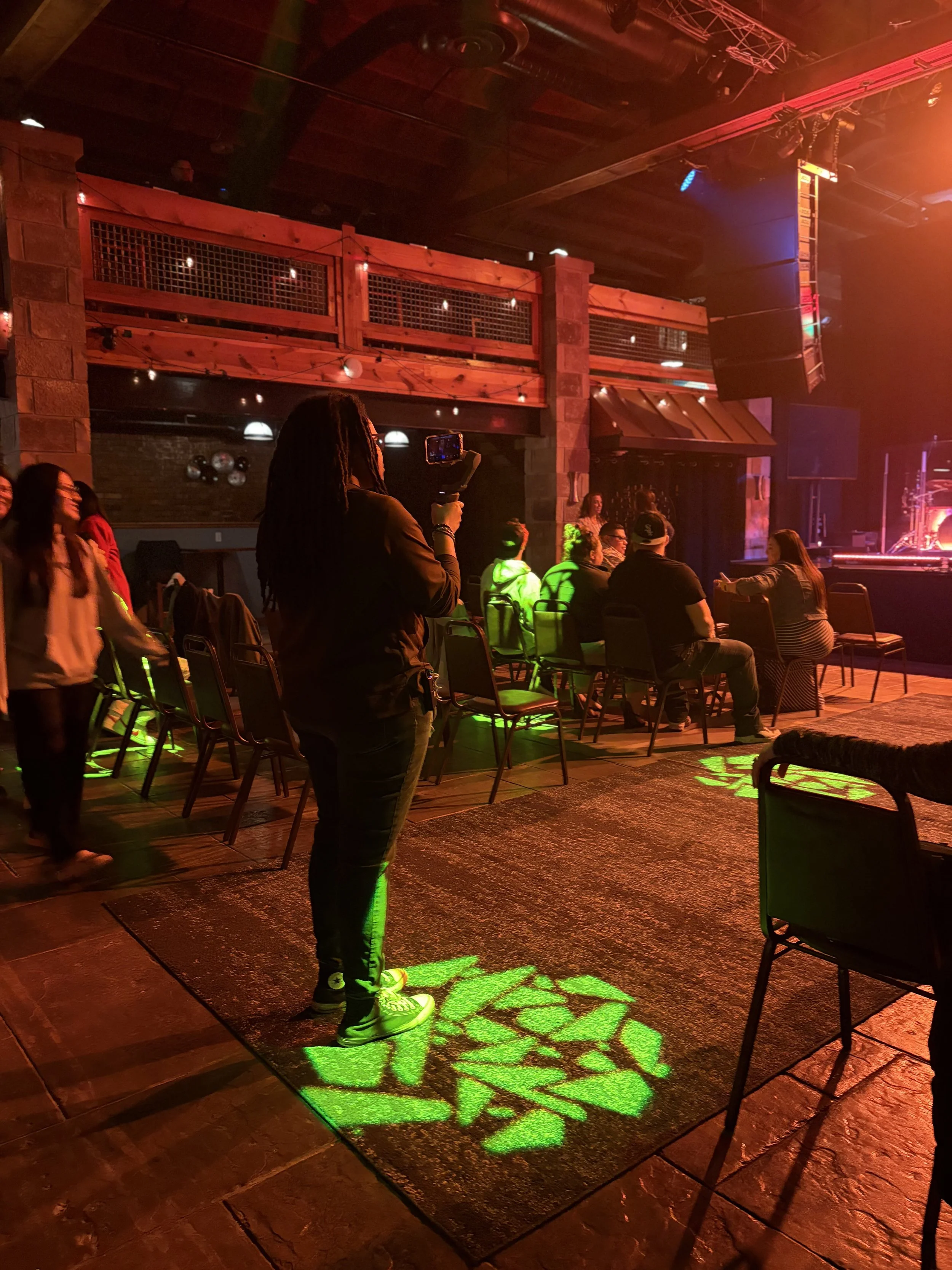 People seated and standing in a dimly lit venue with stage lighting, a woman is taking a photo or video with her phone, and a green leaf-shaped light projection is visible on the floor.