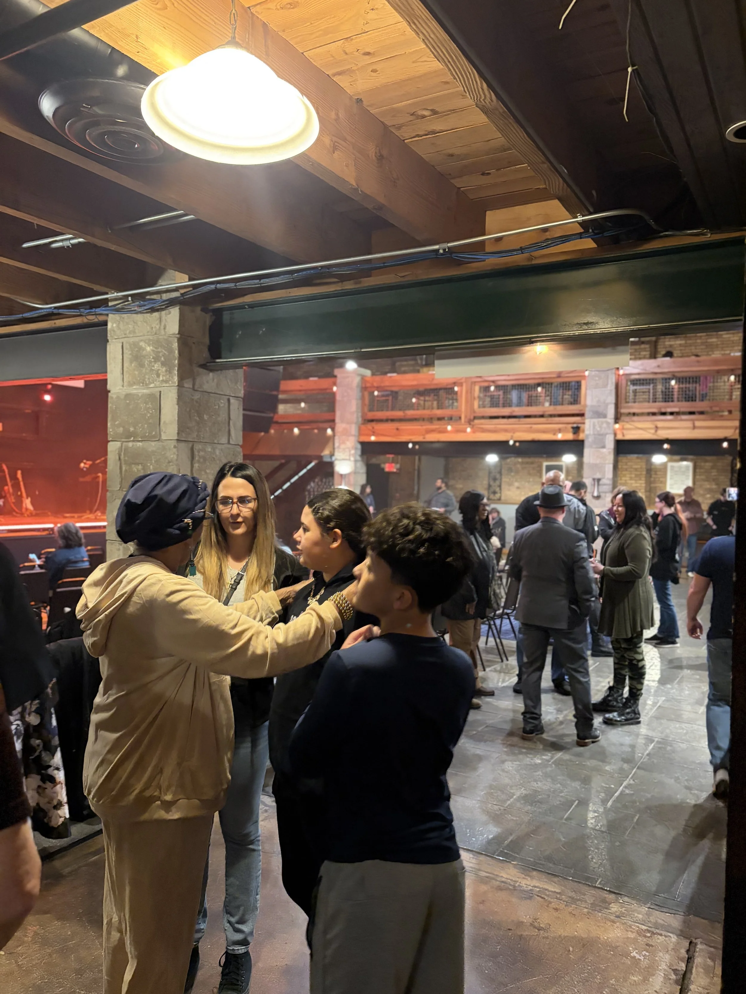 People socializing in an indoor venue with wooden beams and stone pillars, with some sitting and others standing, including a woman in a tan jacket and glasses.