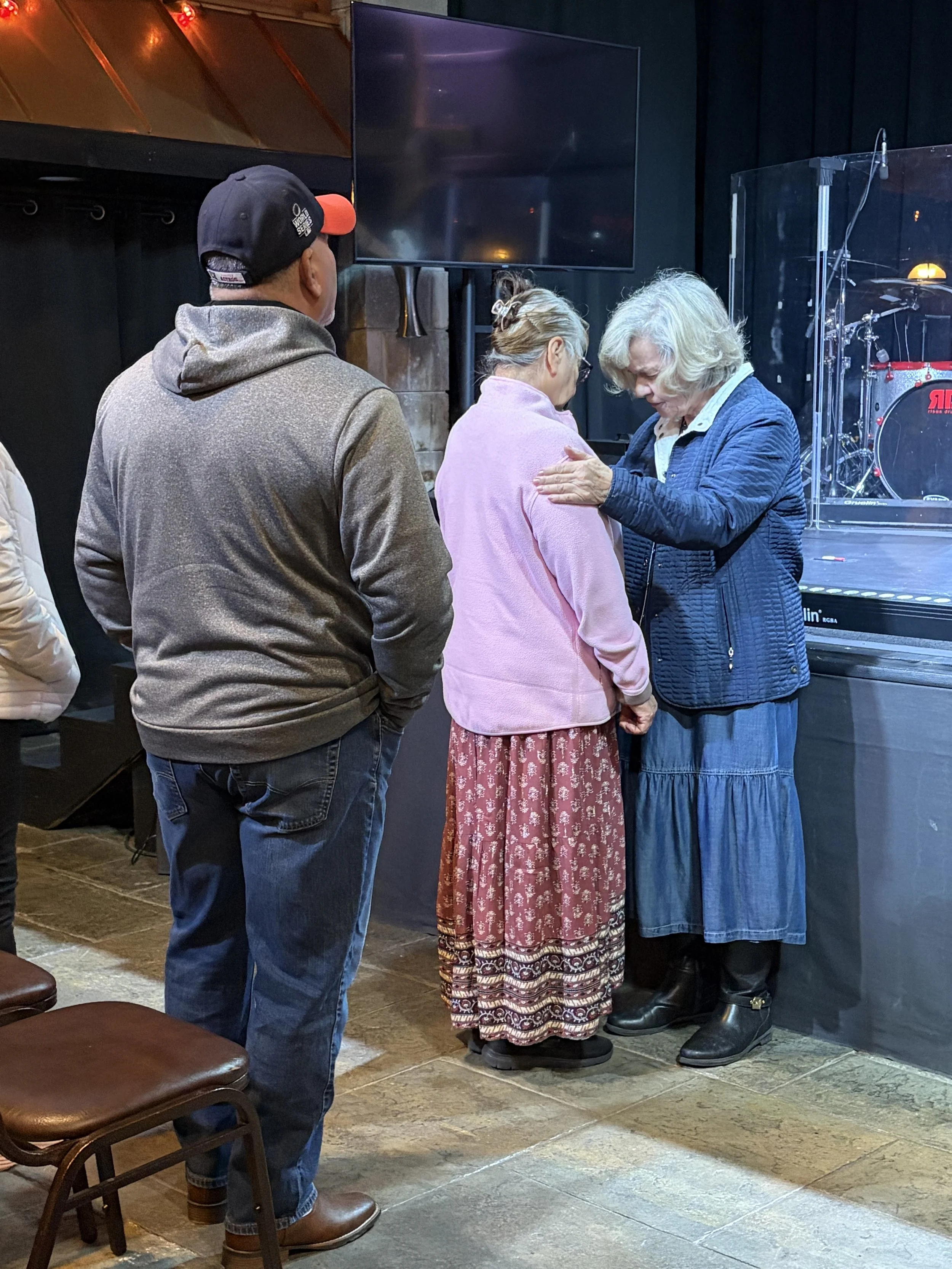 An elderly woman in a pink sweater and patterned skirt is praying with her head bowed and hand on her chest in front of another elderly woman in a blue jacket and long skirt. A man in a hoodie and baseball cap is standing nearby, observing. They are inside a church or meeting hall with a stage, a drum set, and a monitor screen.