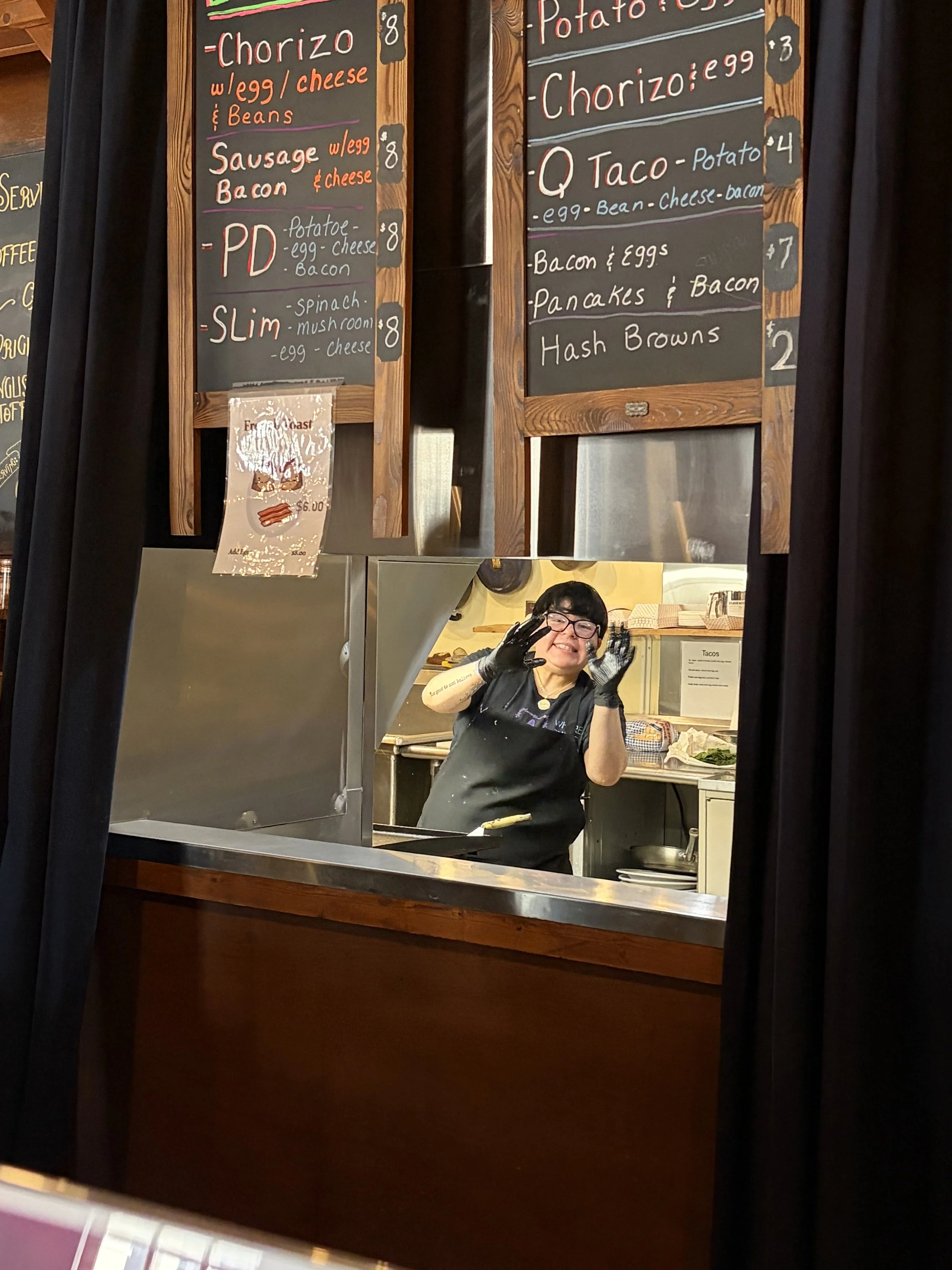 A woman smiling through a serving window, wearing black gloves and a black apron, with dark hair and glasses, behind a food service counter with menu boards overhead.