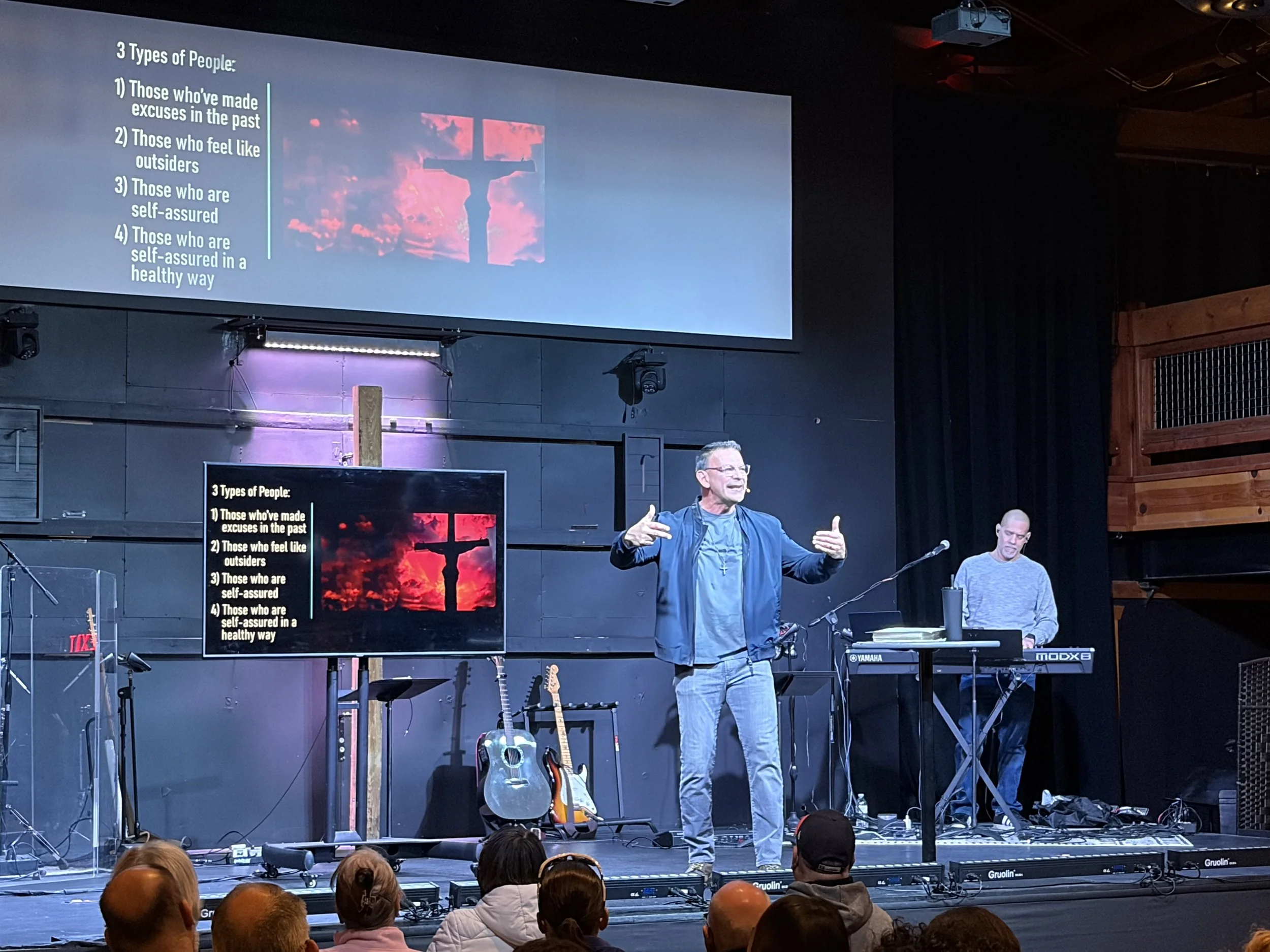 A man is speaking on stage in front of a slide with the text "3 Types of People" and a crash of Jesus on the cross. Several guitars are on stands, and a keyboard is in front of the stage, with an audience watching.