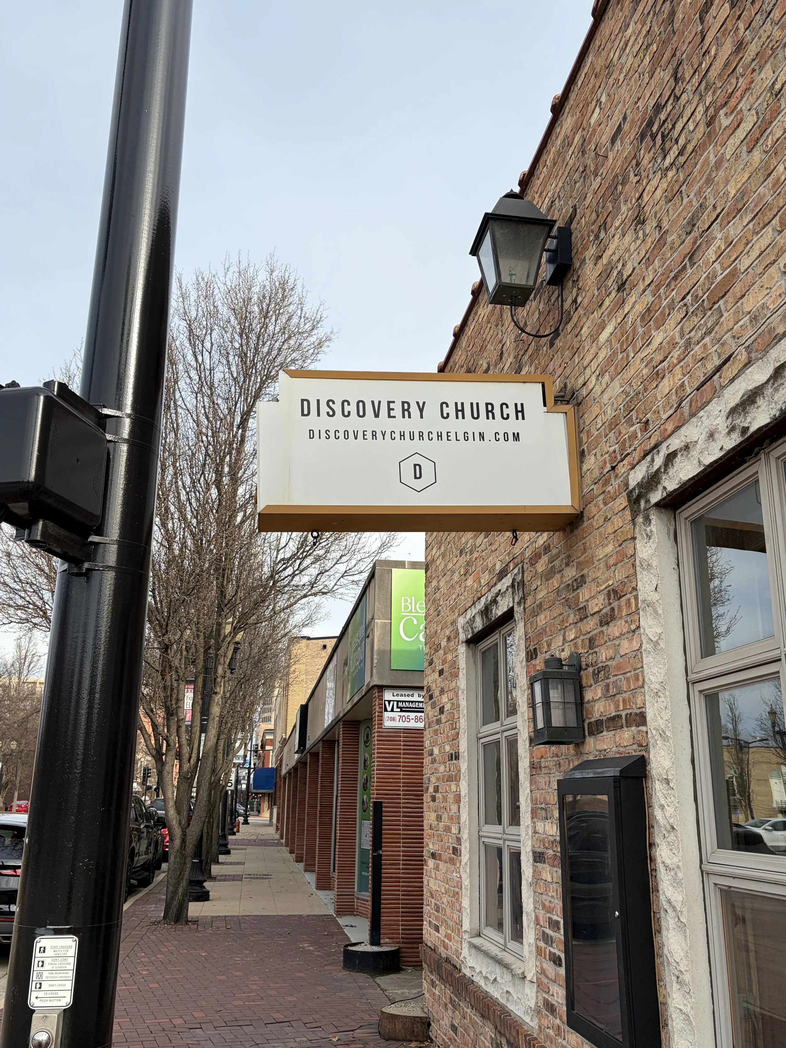 Street view of a brick building with a sign for Discovery Church, a black outdoor lamp, and a parking meter and traffic light pole in the foreground. Leafless trees and storefronts line the sidewalk.