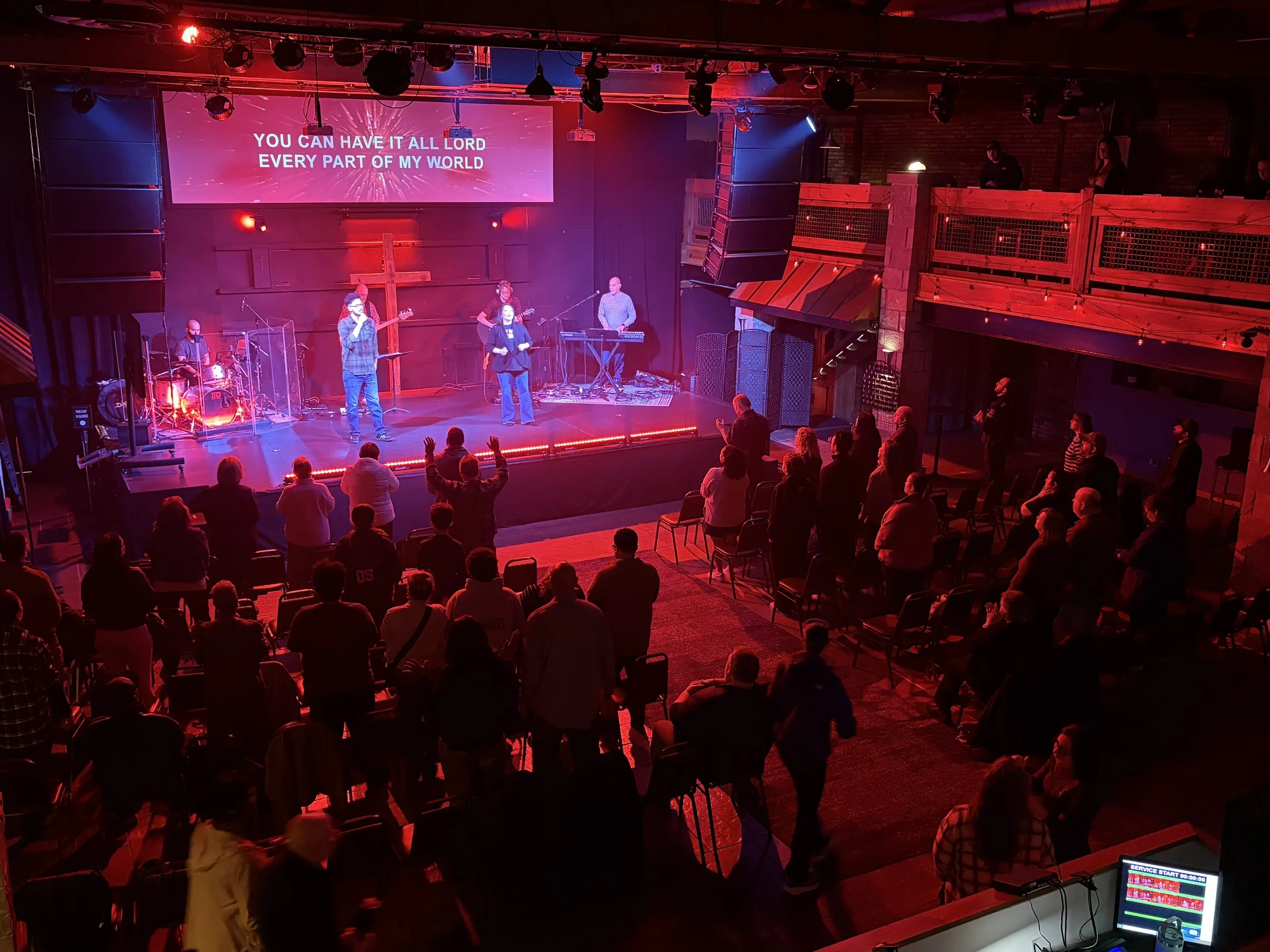A concert stage with a band performing, audience members in front, and a large screen displaying song lyrics. The venue has a rustic interior with red lighting and a balcony with people watching from above.