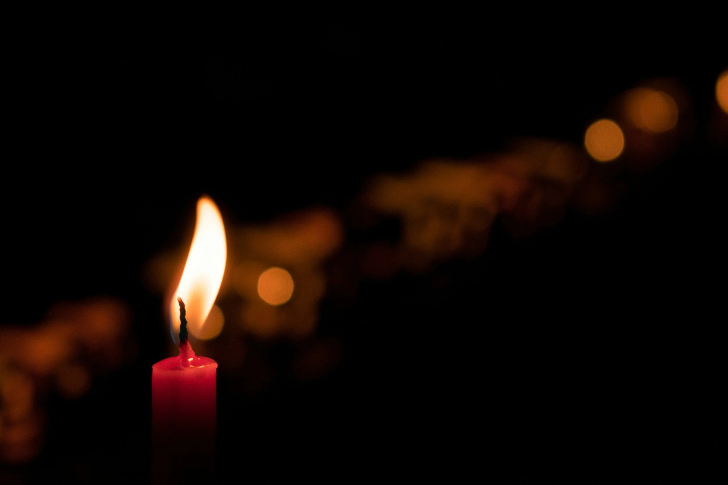 A lit pink candle with a small flame against a dark background, with out of focus orange and yellow lights in the distance.