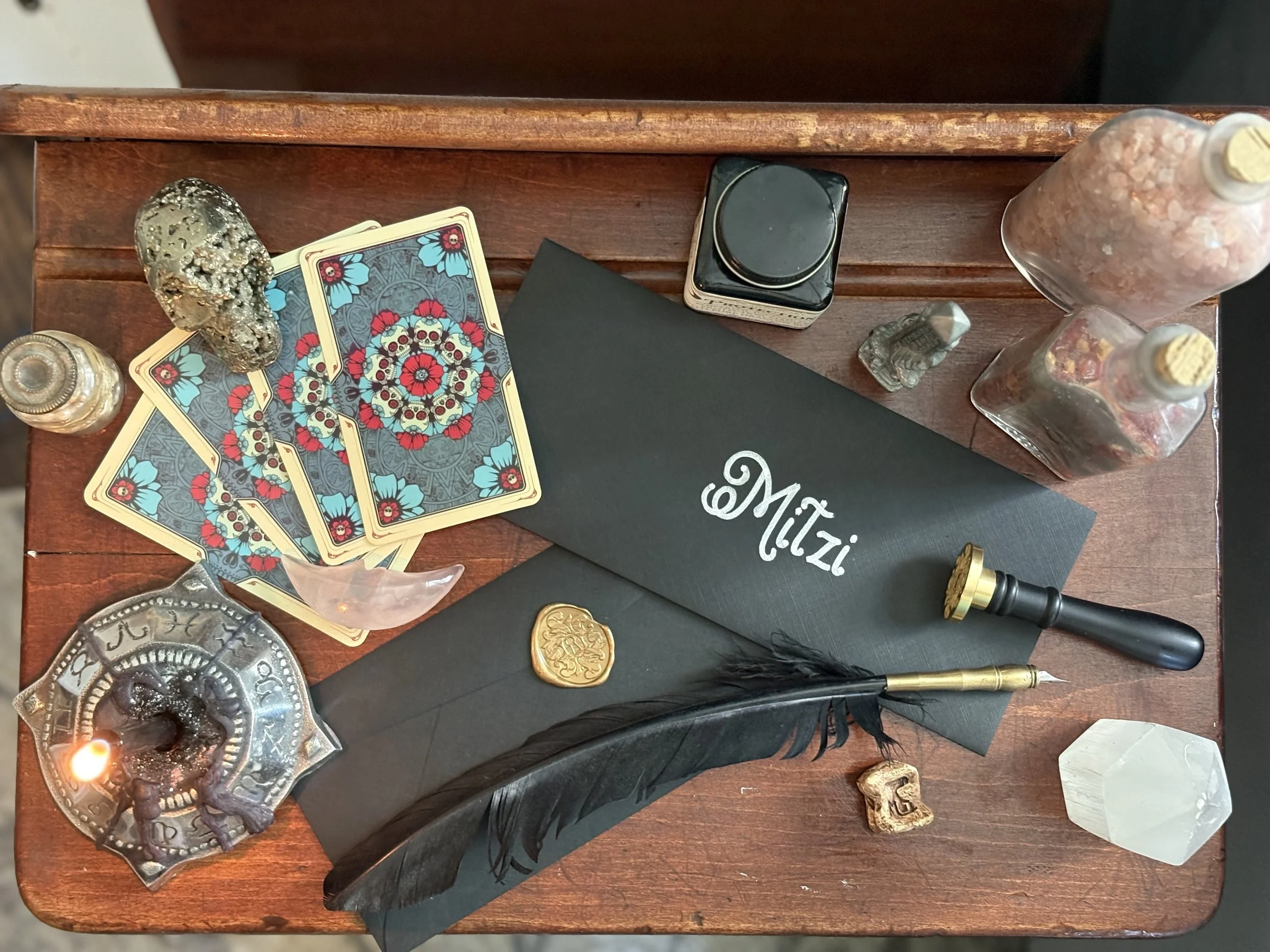 A tarot card reading setup on a wooden table with tarot cards, coins, a black candle, a black notebook labeled 'Mitzzi,' crystals, a skull-shaped incense holder with incense, and decorative bottles and objects.