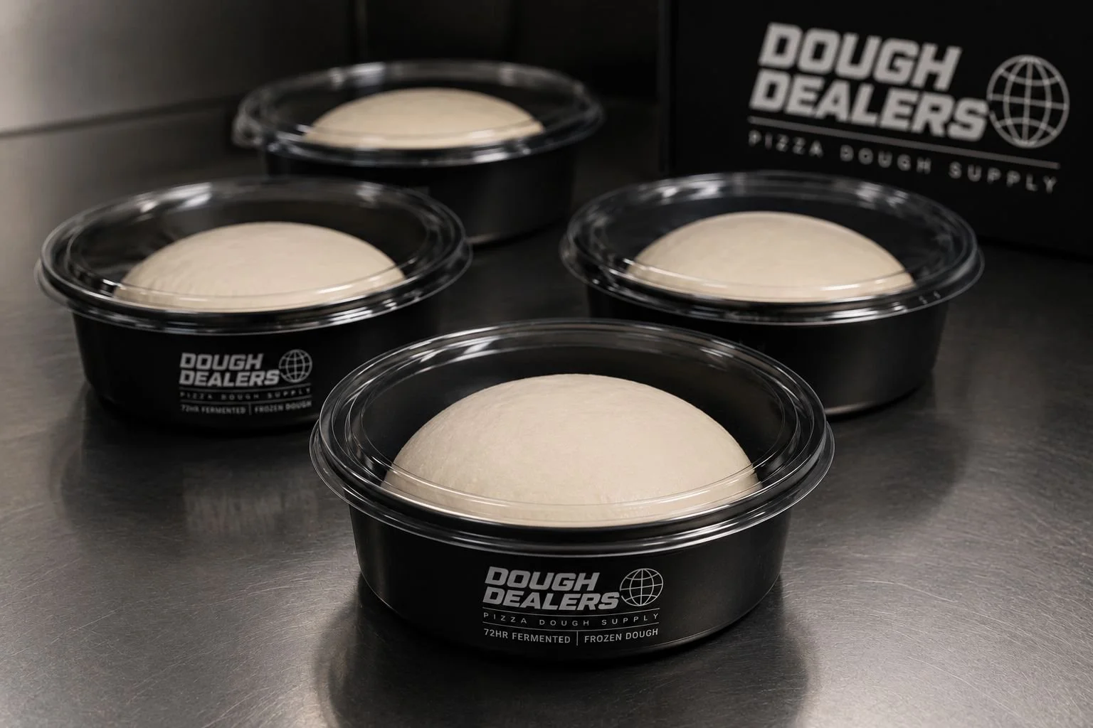 Four black containers with clear lids containing frozen pizza dough from Dough Dealership, with a Dough Dealership box in the background.