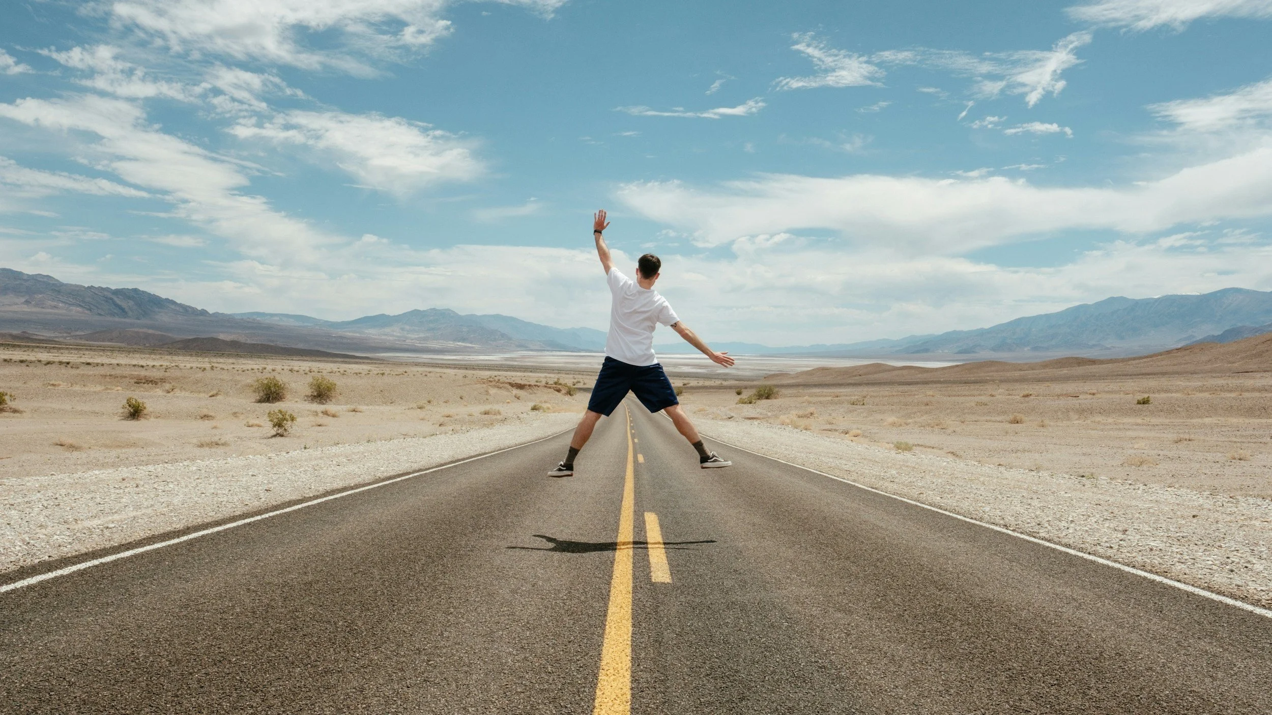 Person standing with arms outstretched in the middle of a deserted road in a desert landscape with mountains and a partly cloudy sky in the background.